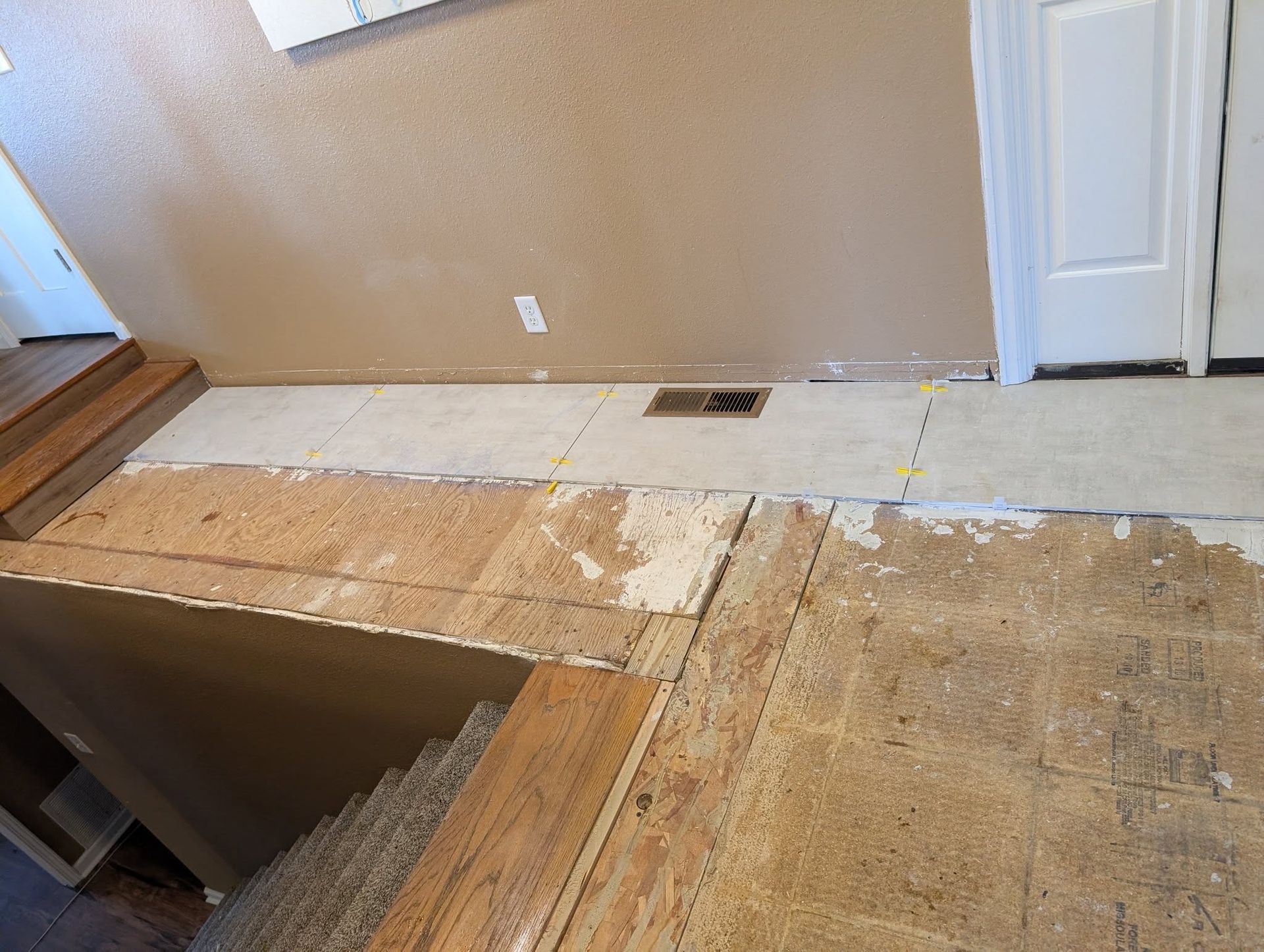 Damaged plywood floor with a large opening and exposed stairs beside a beige wall and white door