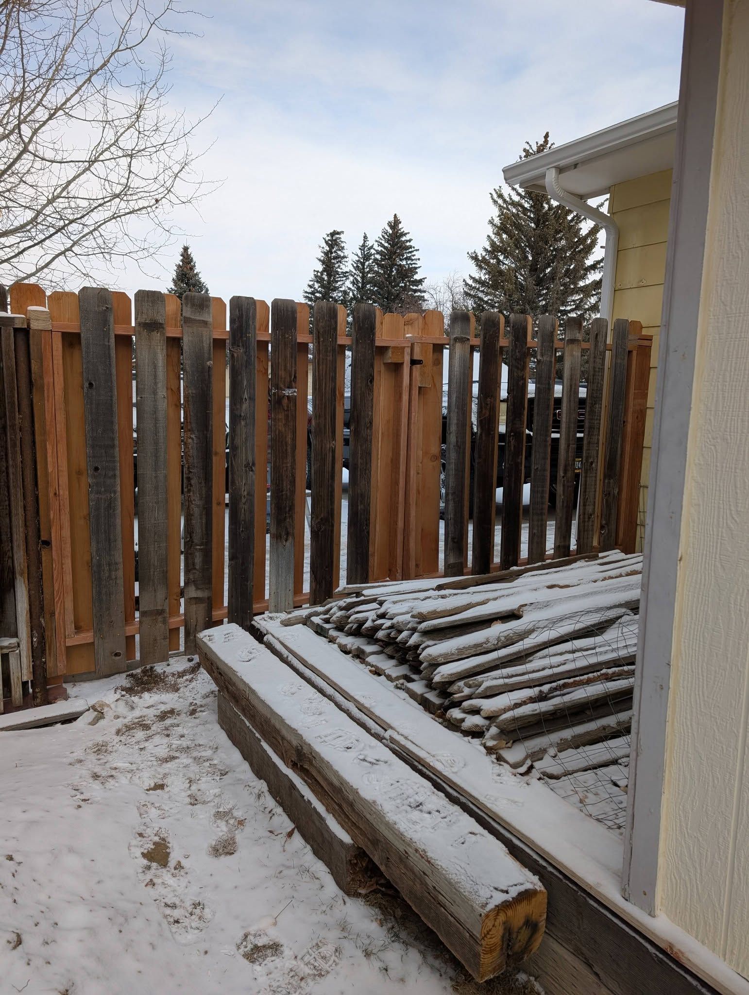 Snow-dusted backyard with wooden fence and stacked boards beside a house.