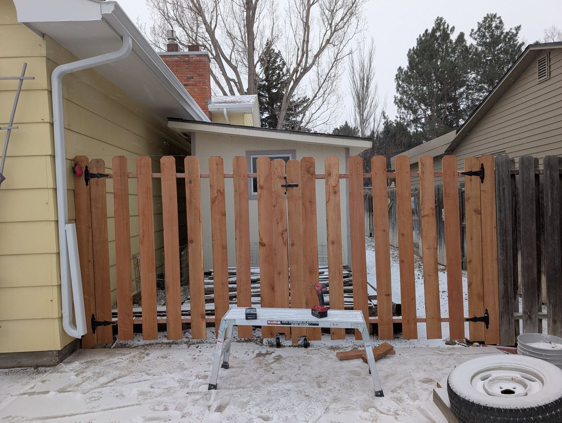 Snowy backyard with a wooden fence and small step stool in front of a yellow house