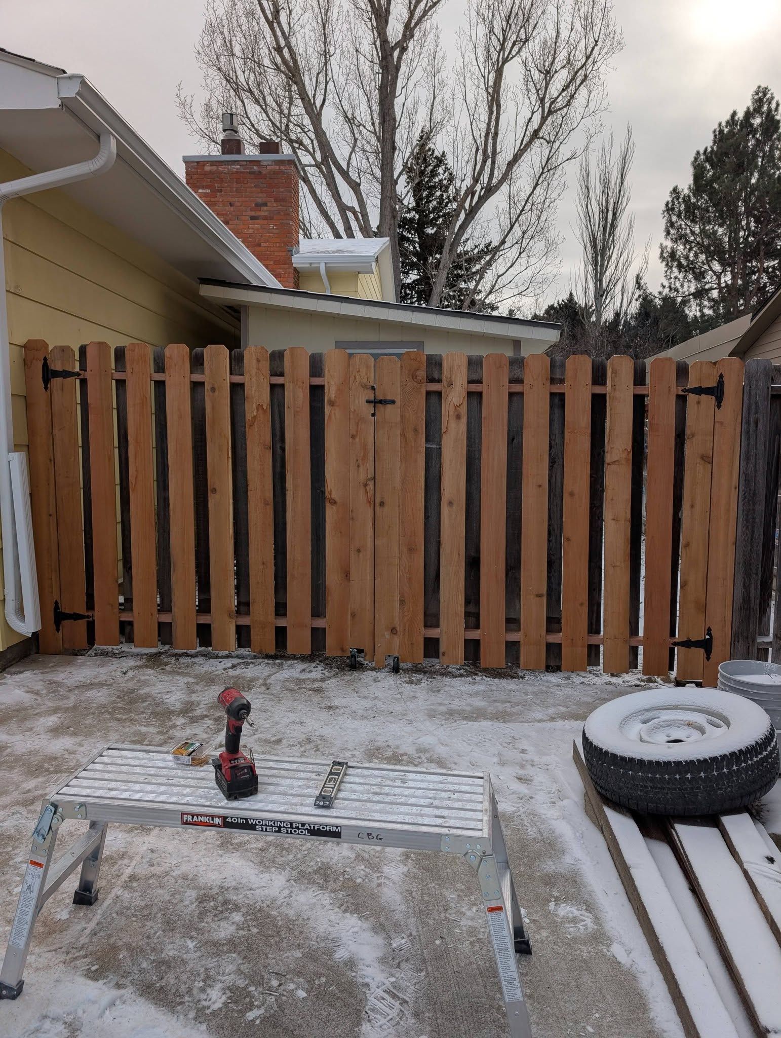 Snowy backyard with a wooden fence, house roof, and outdoor items on a patio.