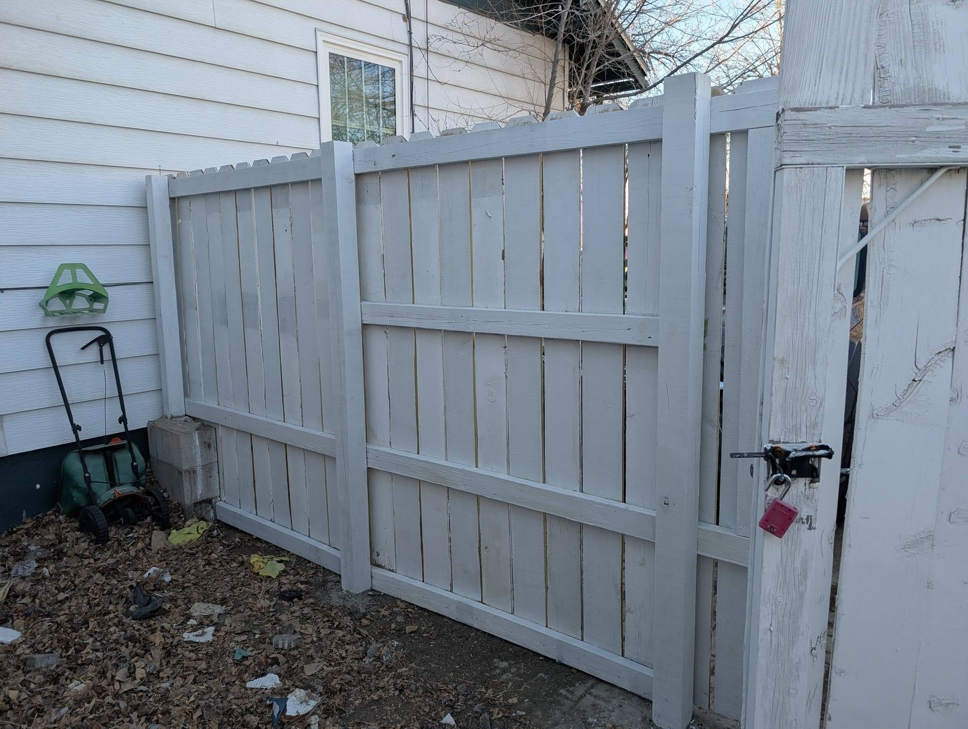 White wooden fence and gate beside a house, with a small green bucket and leaf-covered ground.