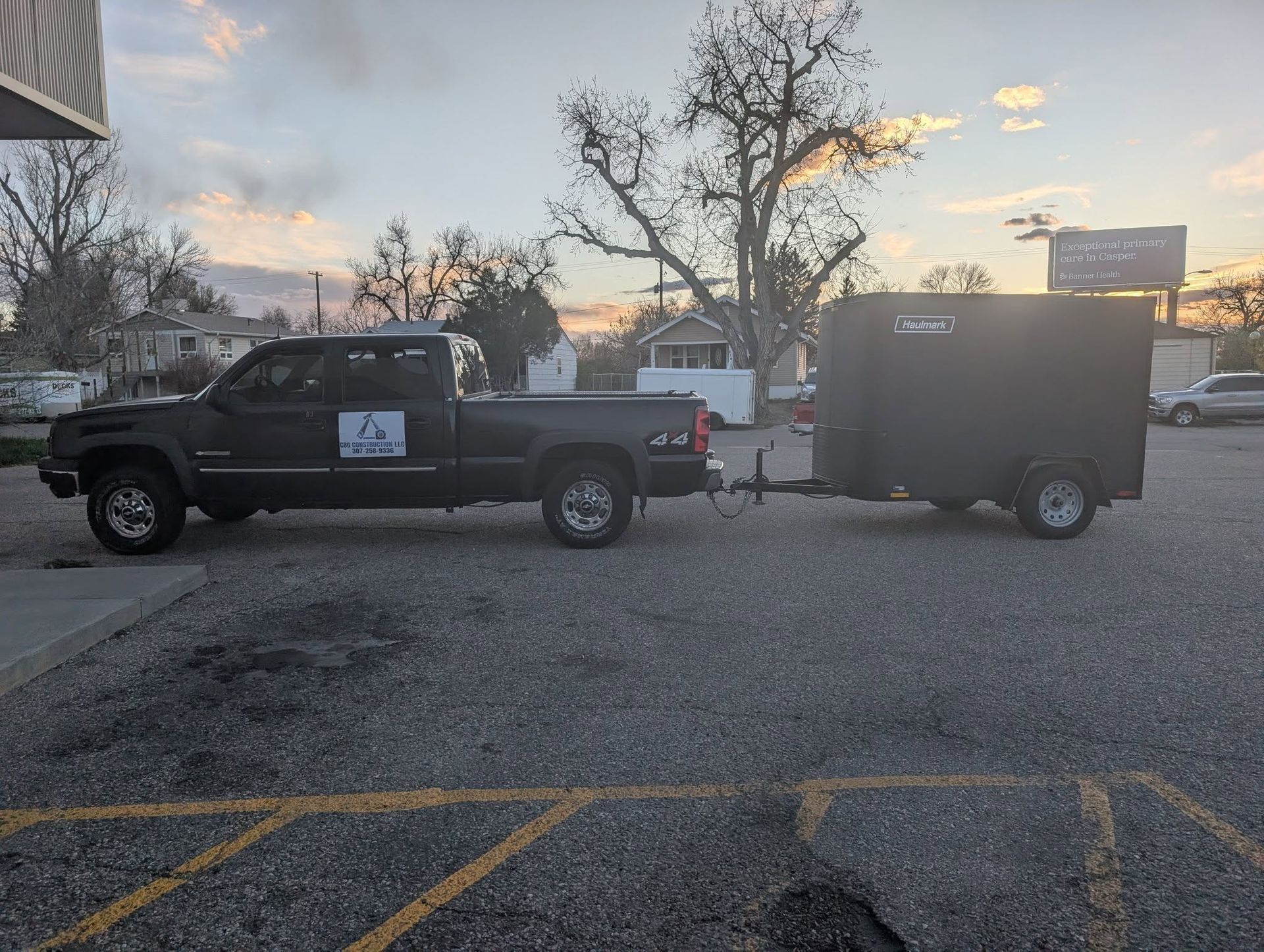 Pickup truck with enclosed trailer parked in a lot at dusk, with bare trees and a sign in the background
