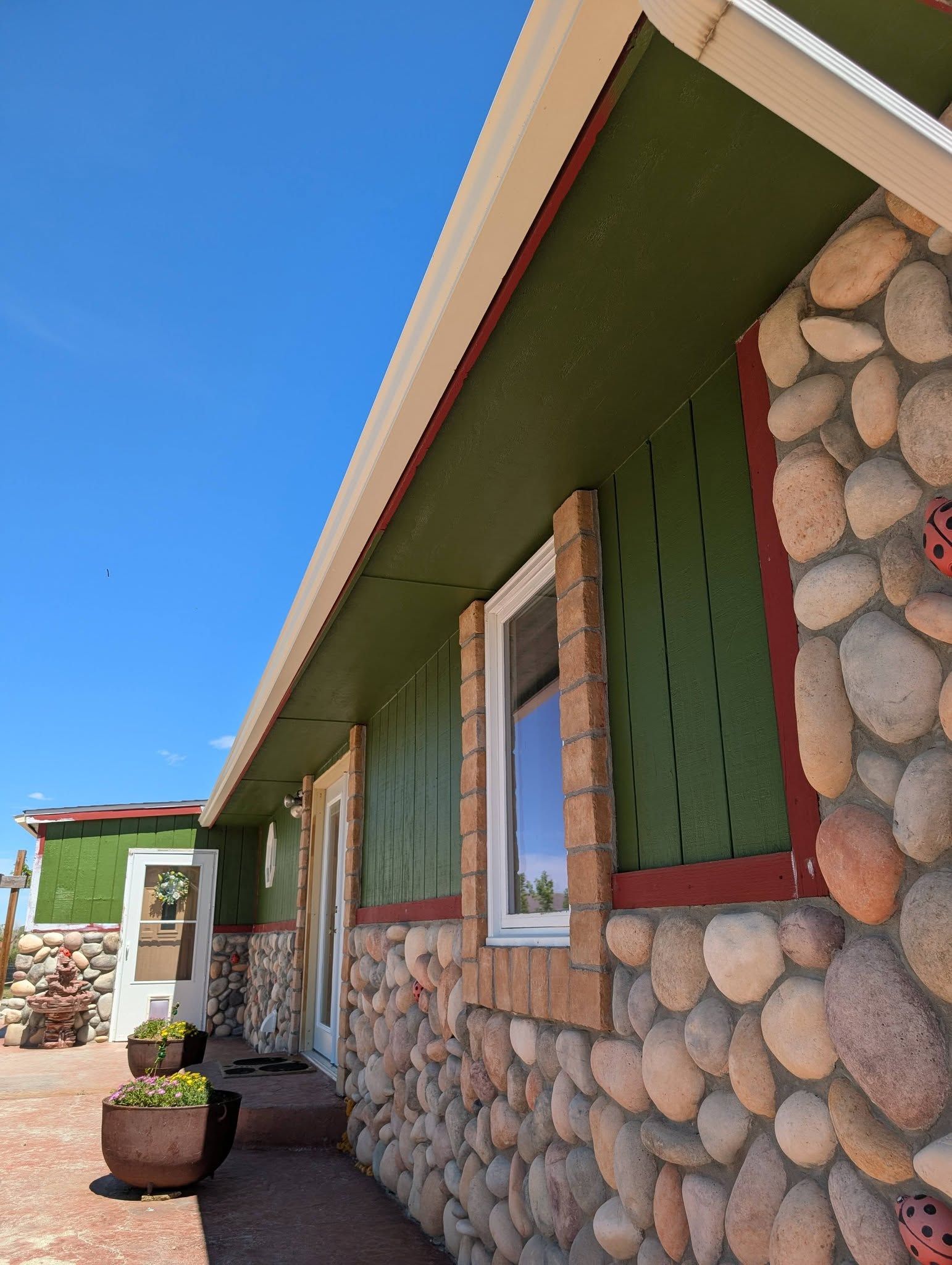 Stone-and-green house exterior with windows, red trim, and a planter under a blue sky.