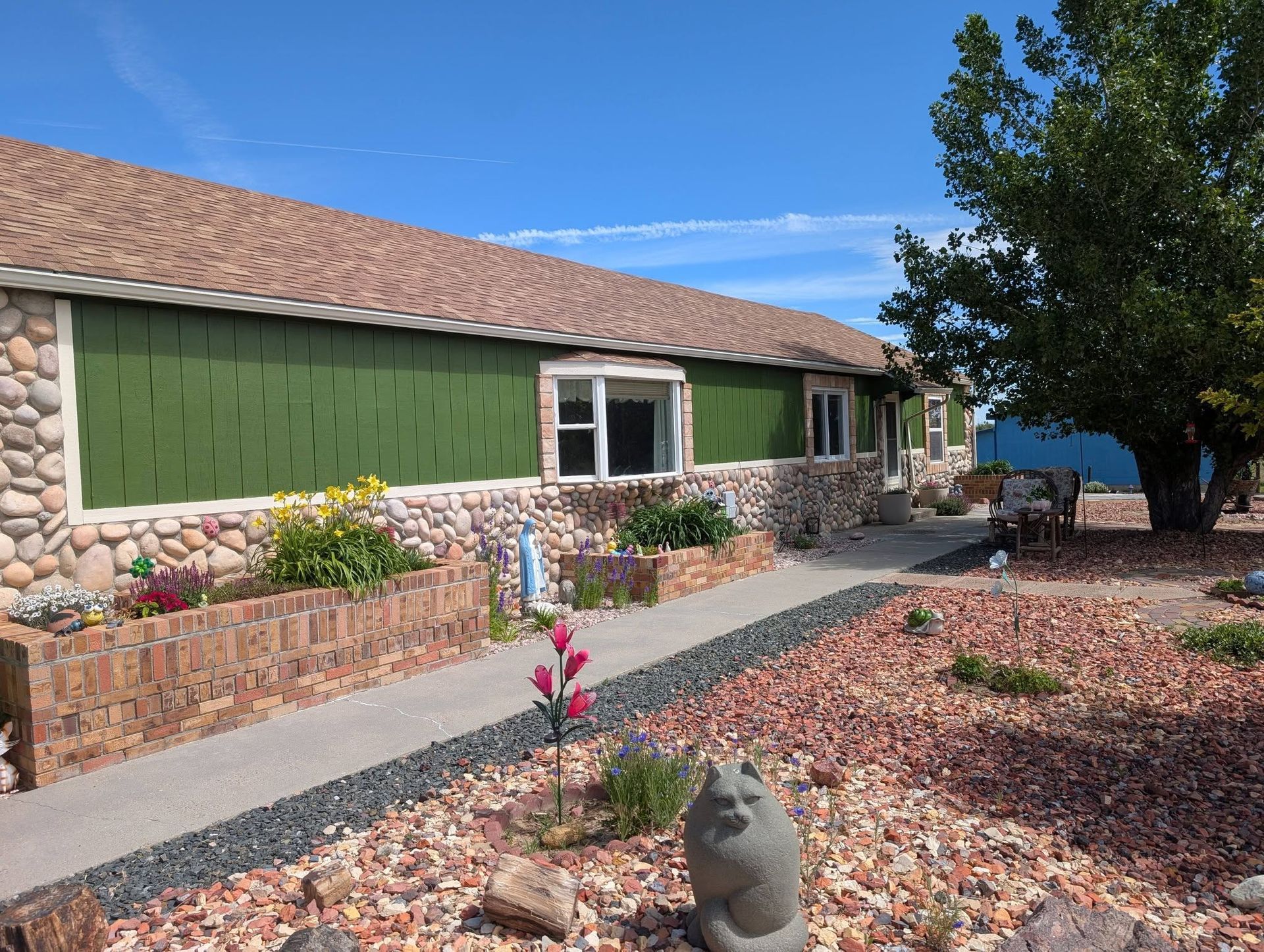 Desert-style house exterior with green siding, stone trim, gravel yard, and a tree under a blue sky
