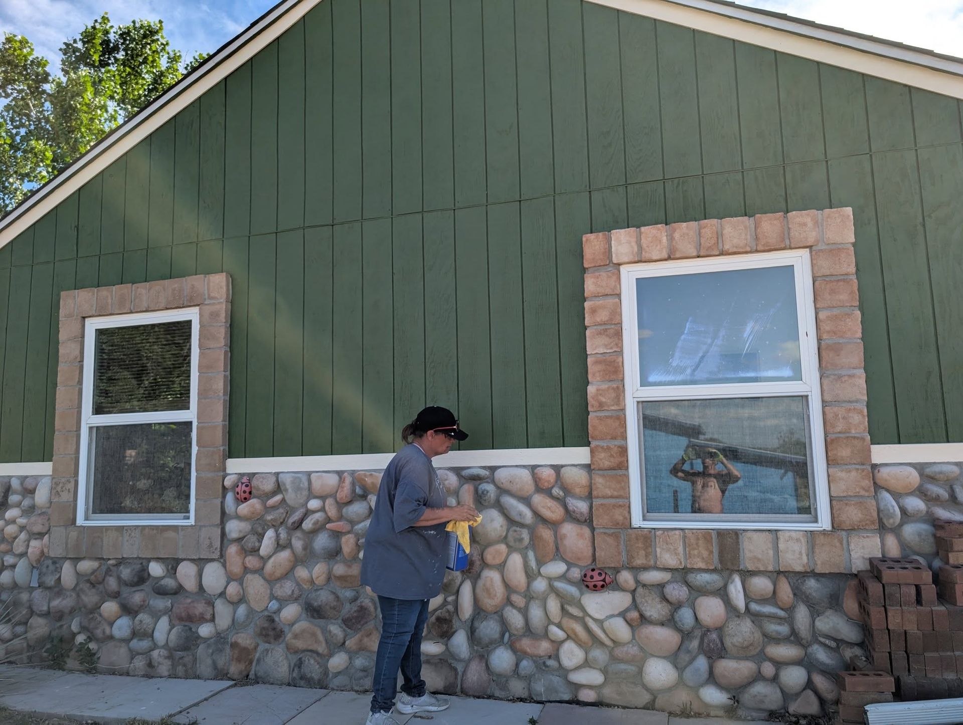 Person painting the side of a green house with stone lower siding and two windows.