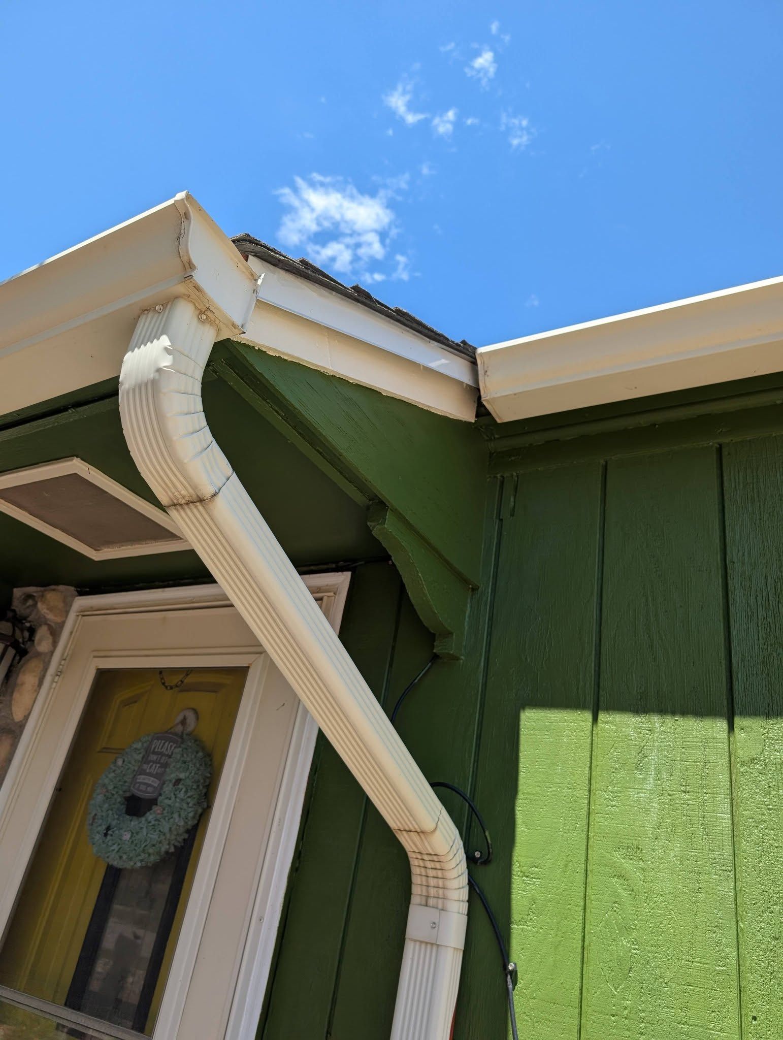 Damaged white rain gutter hanging from a green house roof against a blue sky