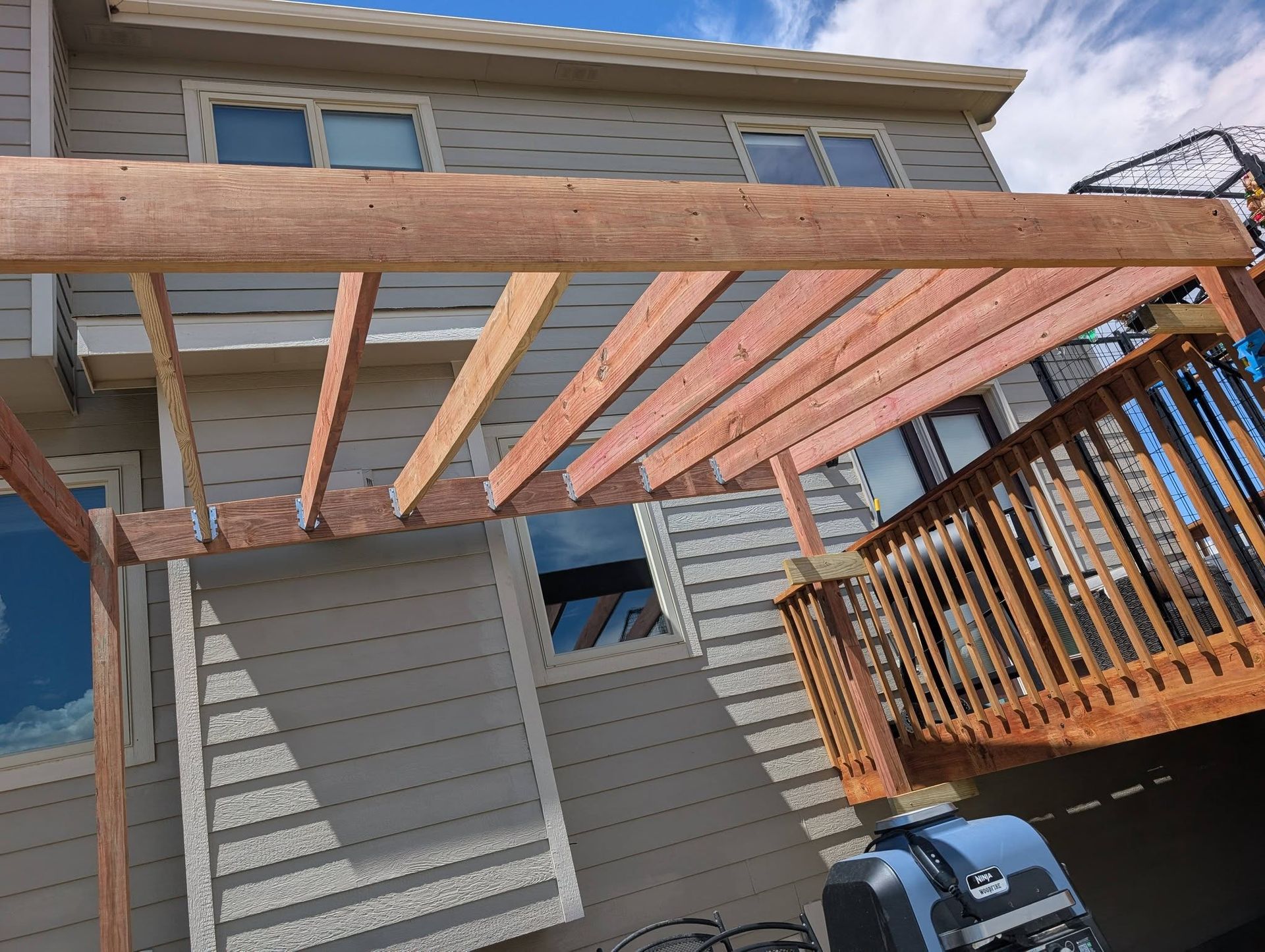 Wooden deck pergola under construction beside a gray house, with a grill on the patio.