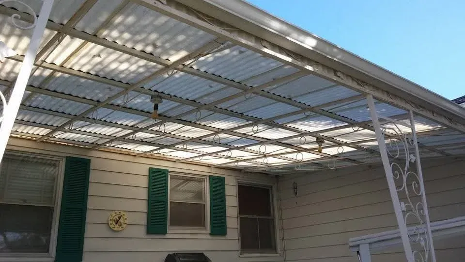 Covered patio with corrugated metal roof, white posts, and green shutters on a beige house
