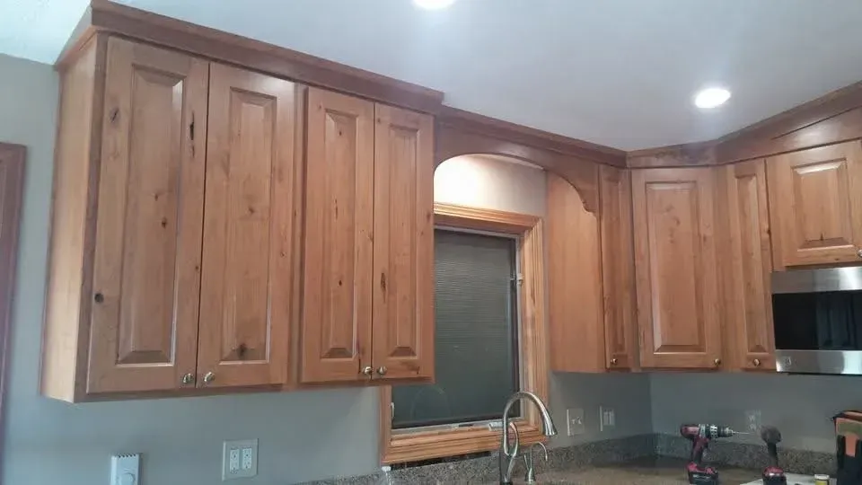 Wooden kitchen cabinets above a sink and window under recessed ceiling lights.
