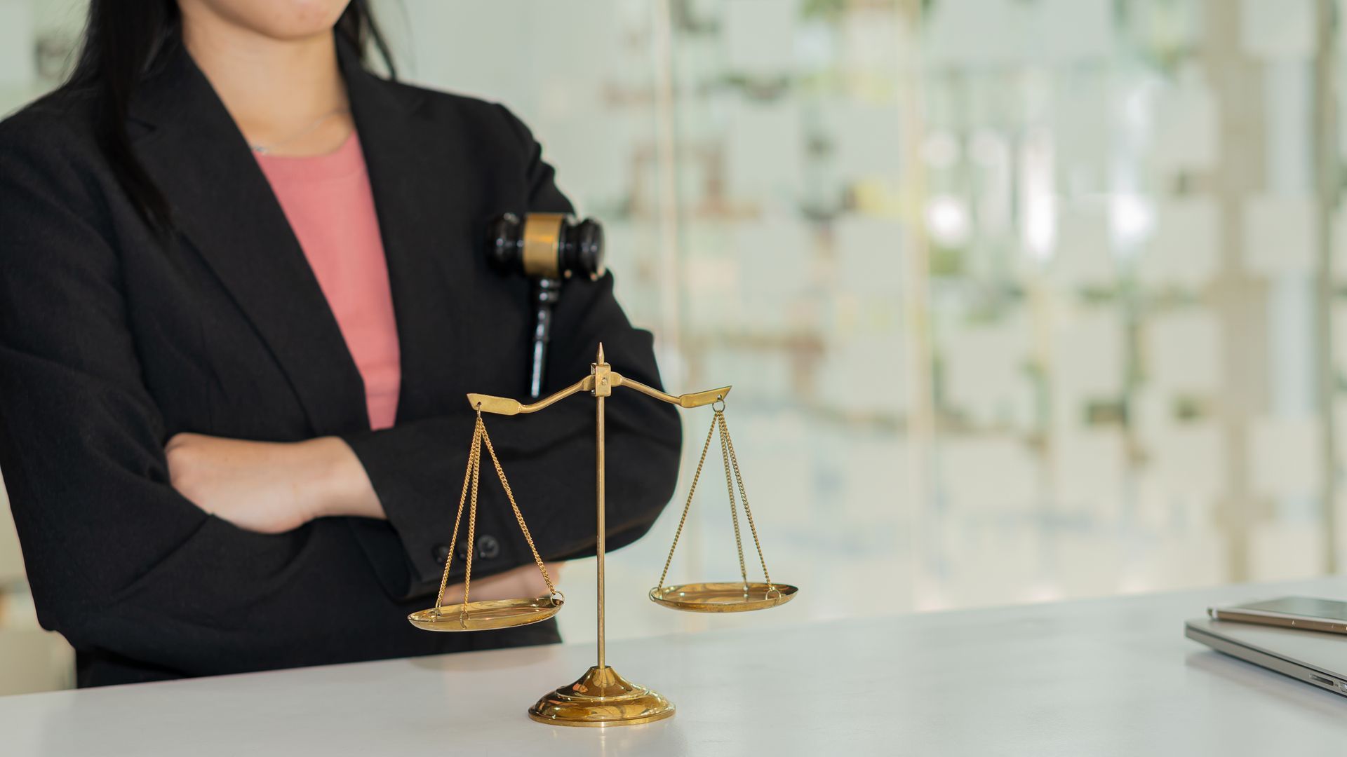 Woman in a blazer with arms crossed, next to a golden scale and gavel on a white table.
