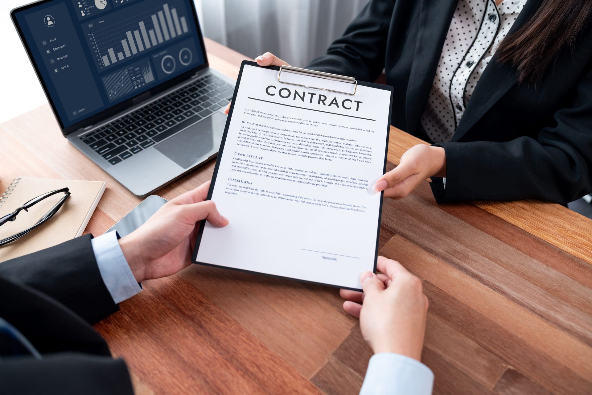 Business meeting with a contract being signed at a wooden desk, with a laptop and pens nearby