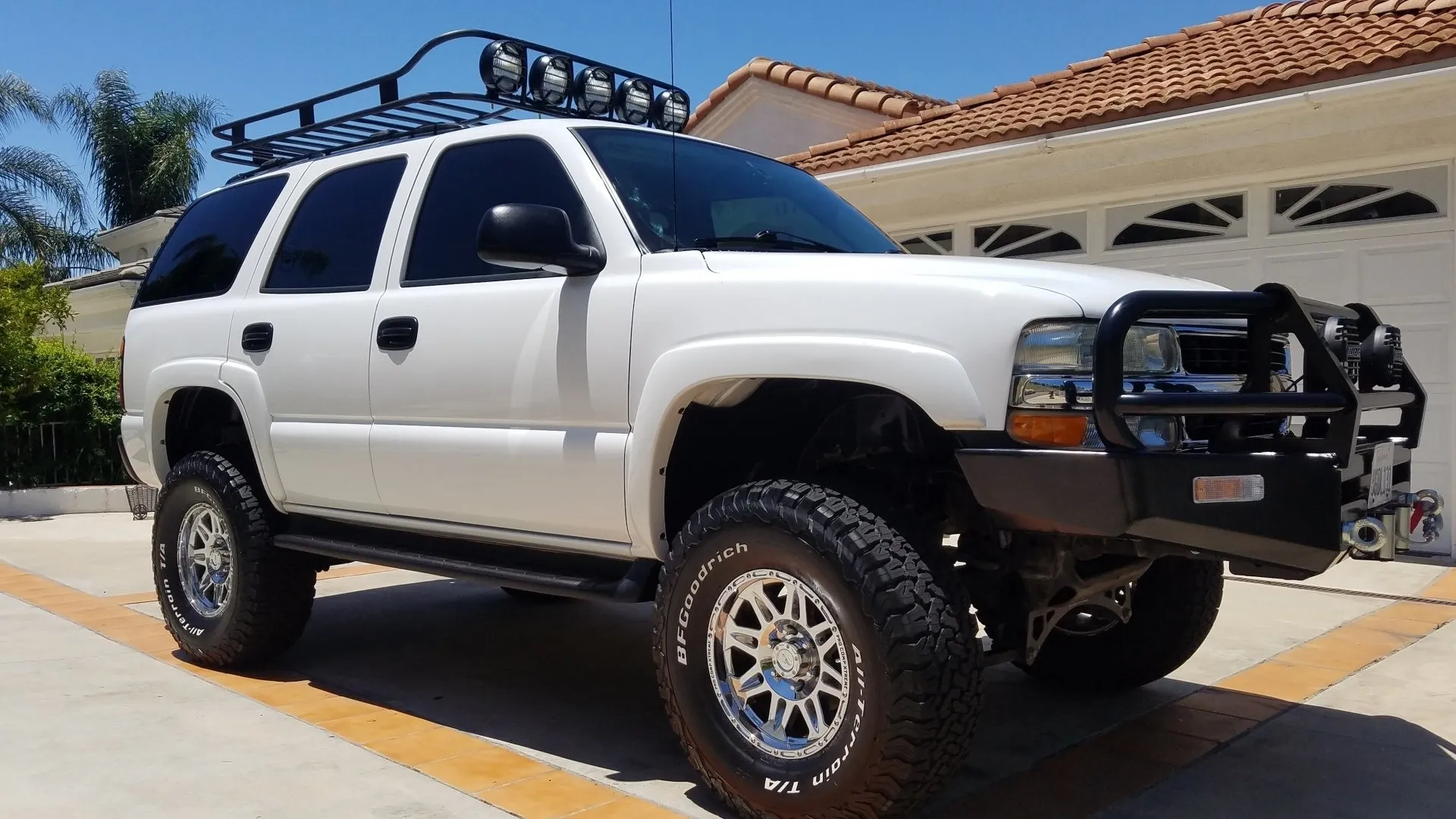 White lifted SUV with black roof rack and front bumper guard parked in driveway.