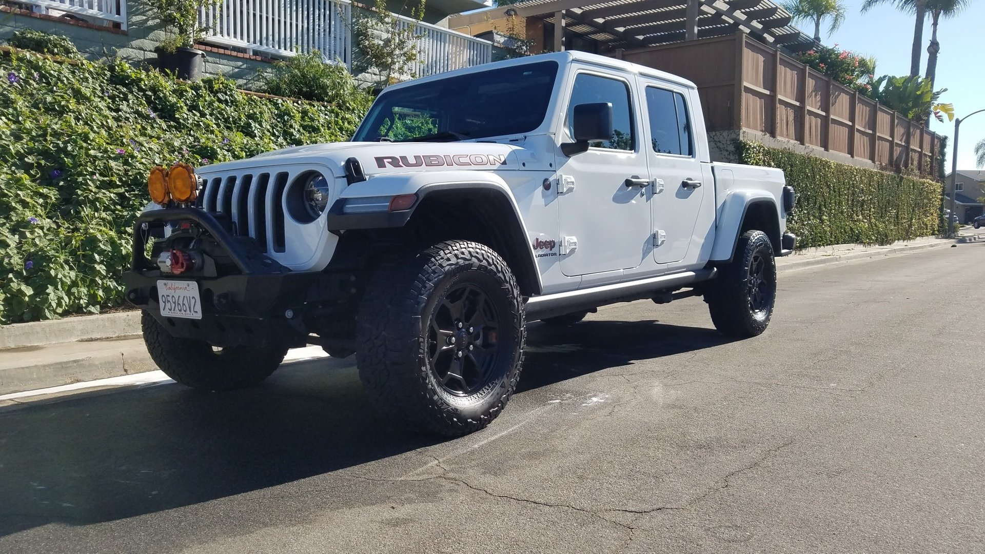 White Jeep Gladiator pickup truck parked on a sunny street, featuring black wheels and off-road accessories.