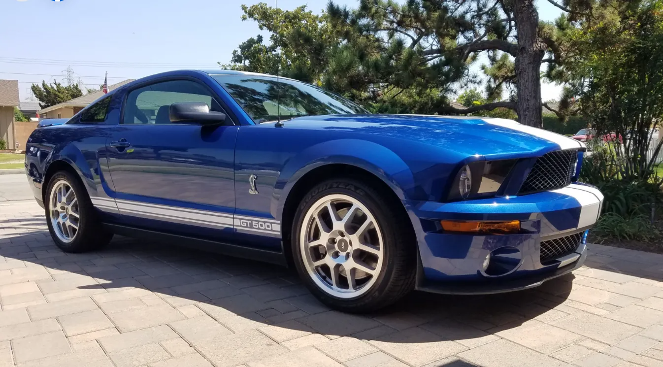 Blue Shelby Mustang coupe parked on a brick driveway, white racing stripes.