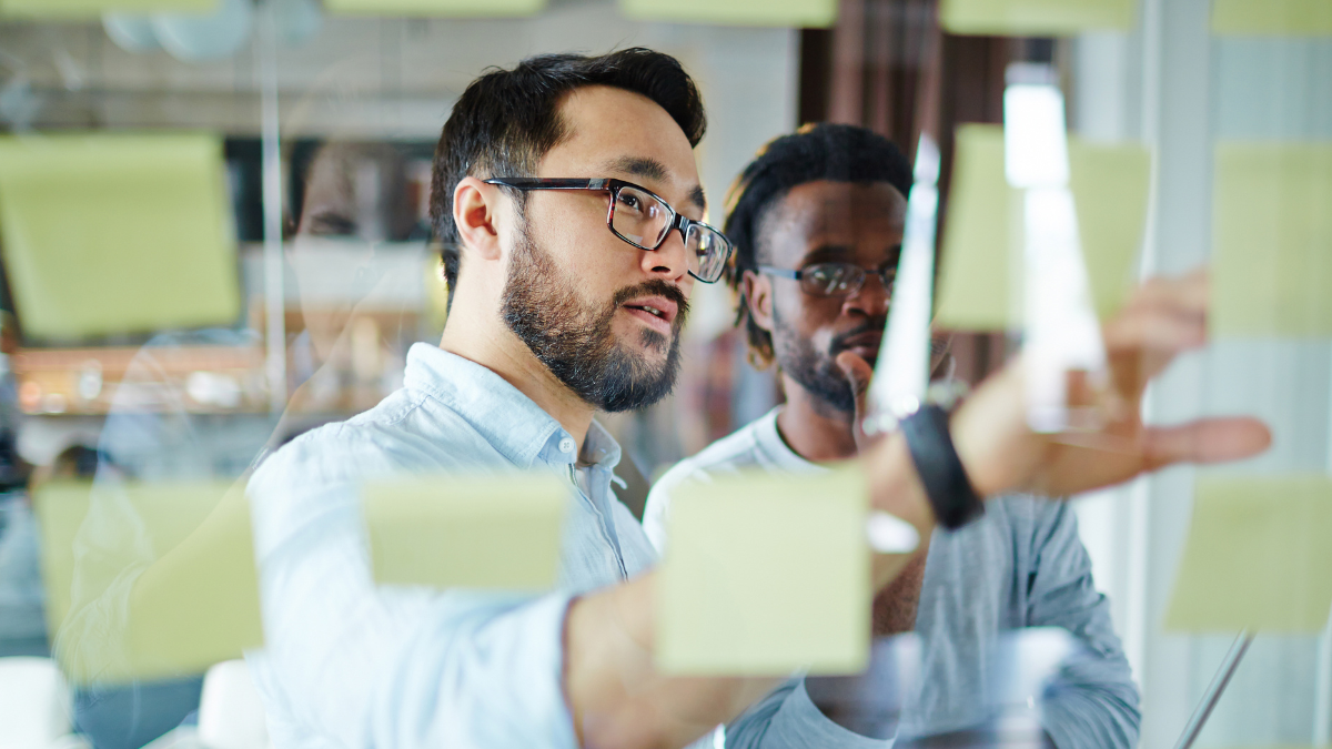 Two men are looking at sticky notes on a glass wall.