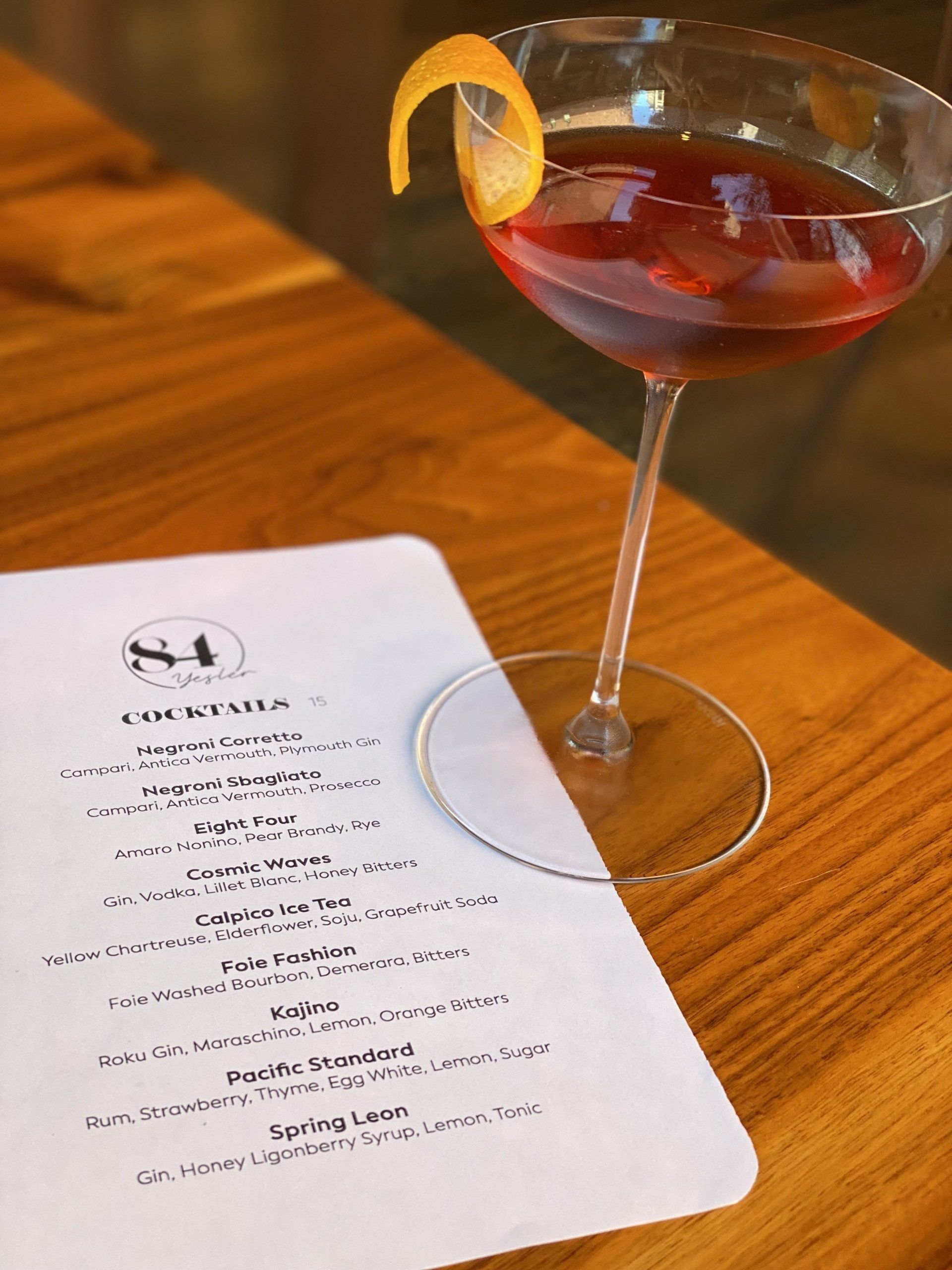 A martini glass with a lemon peel on top of it is on a wooden table next to a menu.