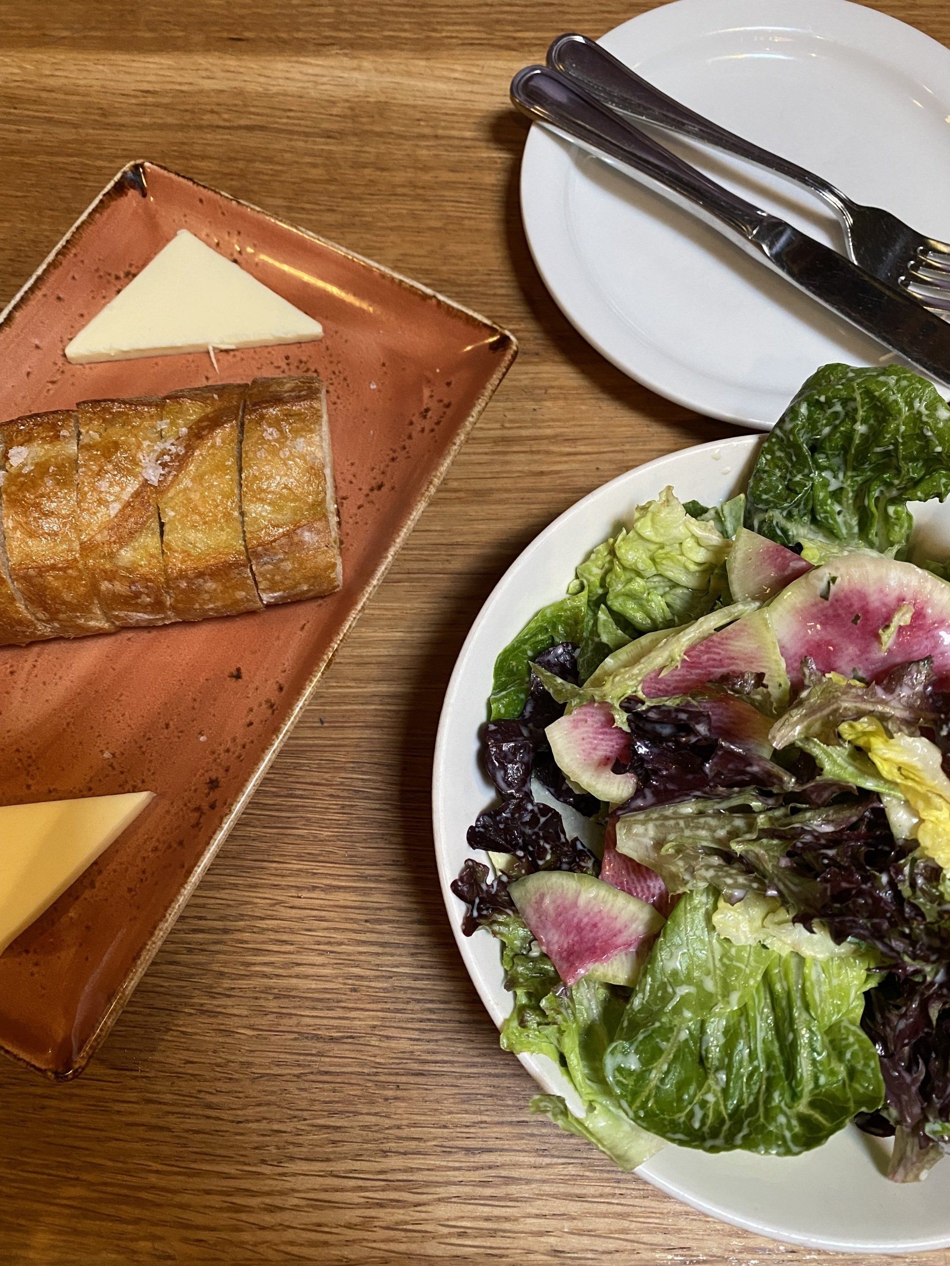 A plate of salad next to a plate of cheese and bread on a wooden table.