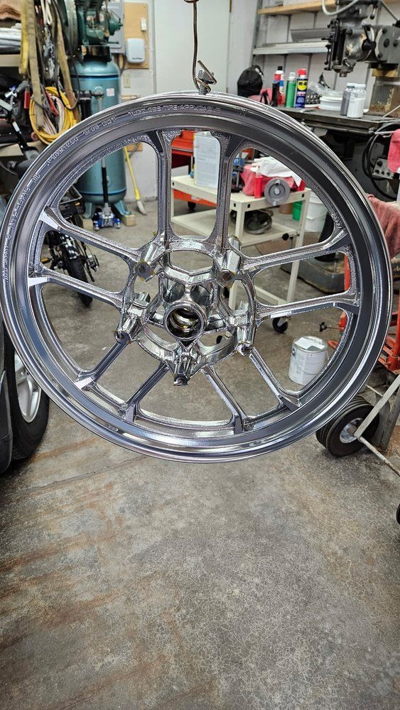 A shiny, chrome-finished custom motorcycle wheel with a multi-spoke design hanging in a cluttered workshop.