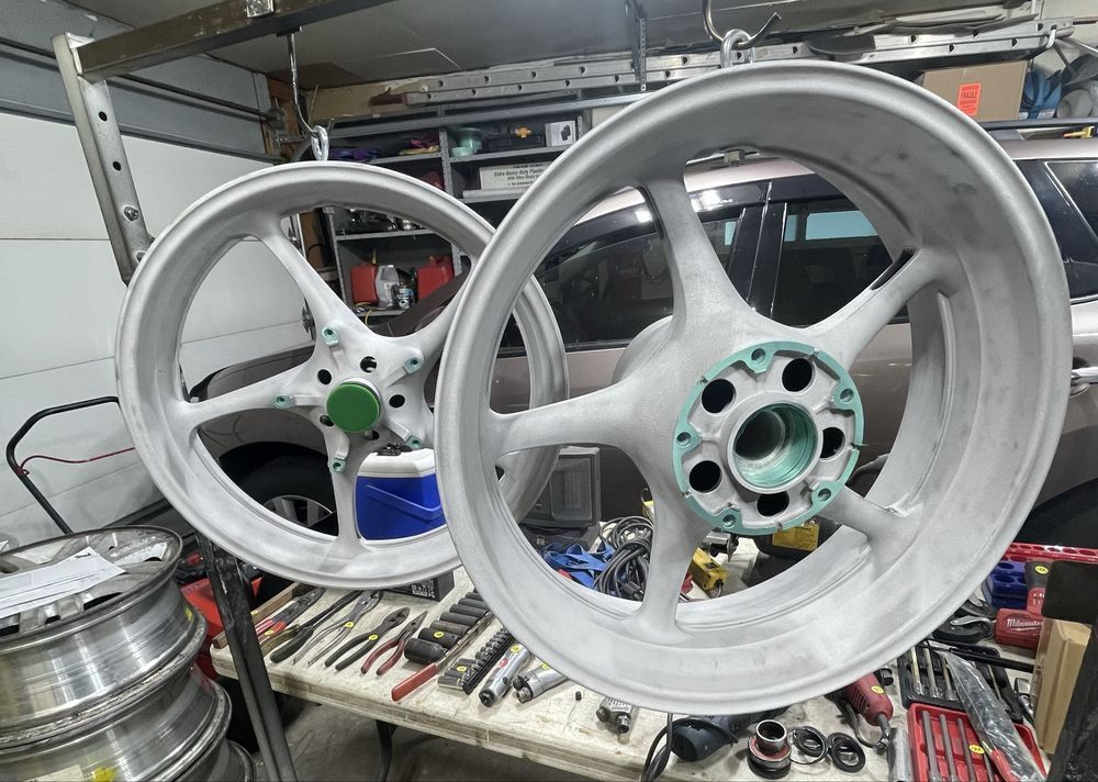 Two light-colored motorcycle wheels suspended from a metal rack in a workshop, with various tools on a table below.