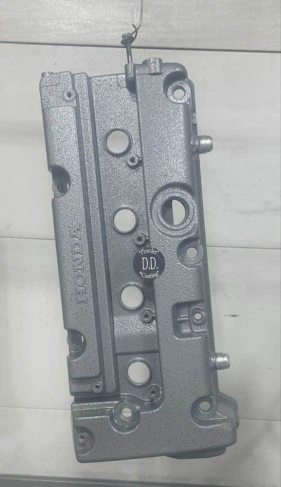A gray, textured Honda valve cover hanging by a wire against a light-colored wooden wall.