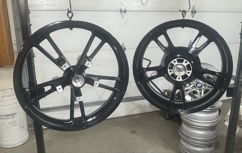 Two black, five-spoke wheels hanging from hooks in a garage, with a stack of silver wheels in the background.