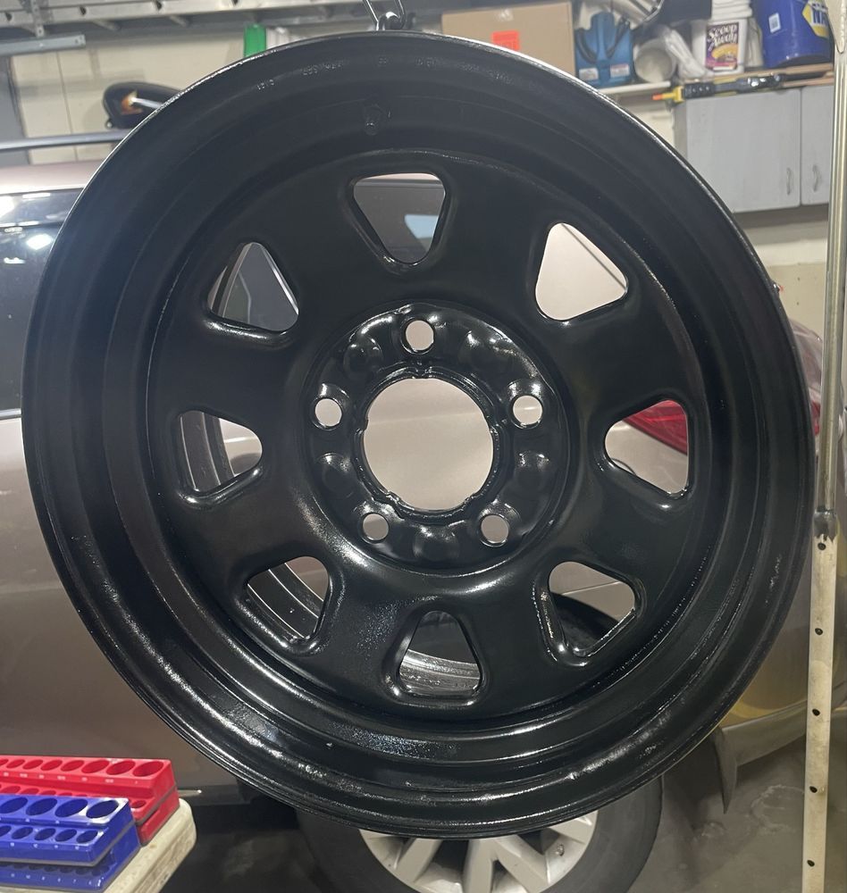 A freshly painted, glossy black automotive steel rim with an eight-hole design, shown in a garage setting.