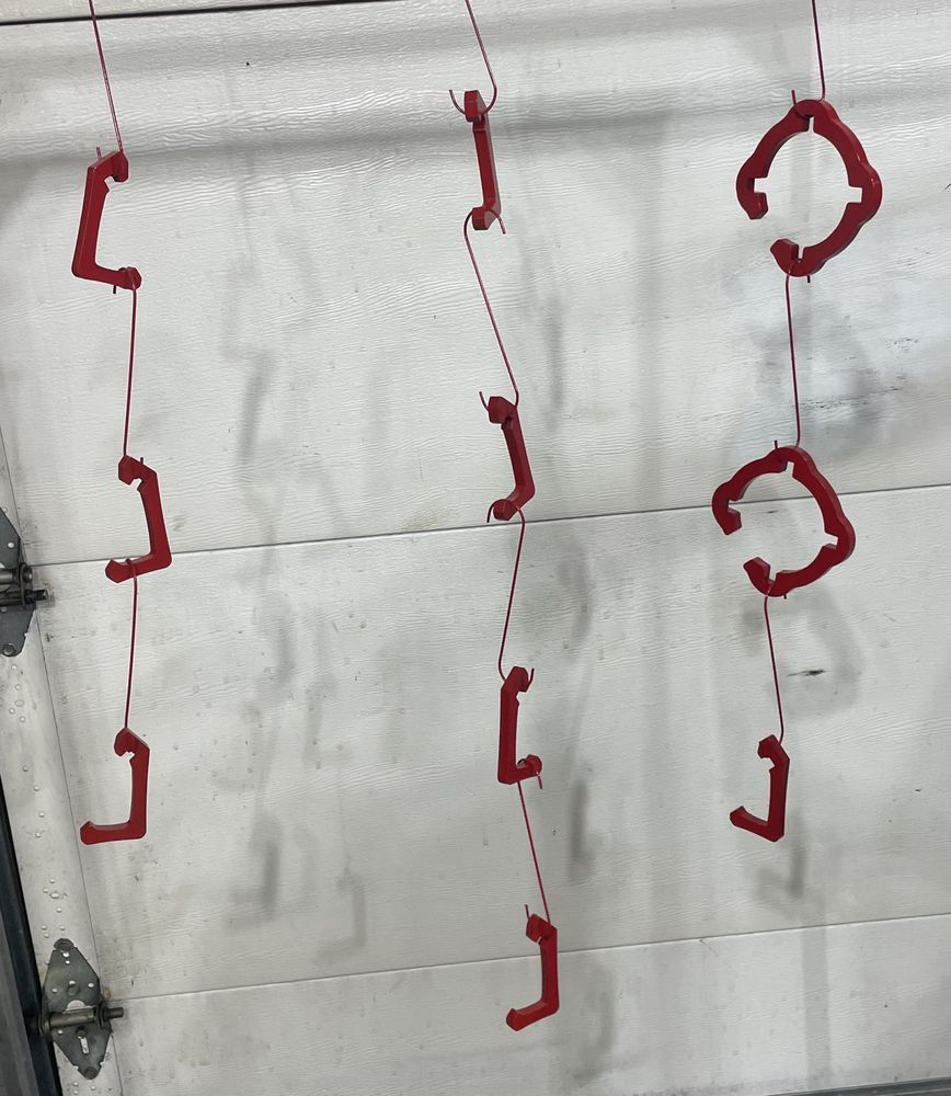 Red, L-shaped and C-shaped metal parts hang by hooks in rows against a white garage door background.