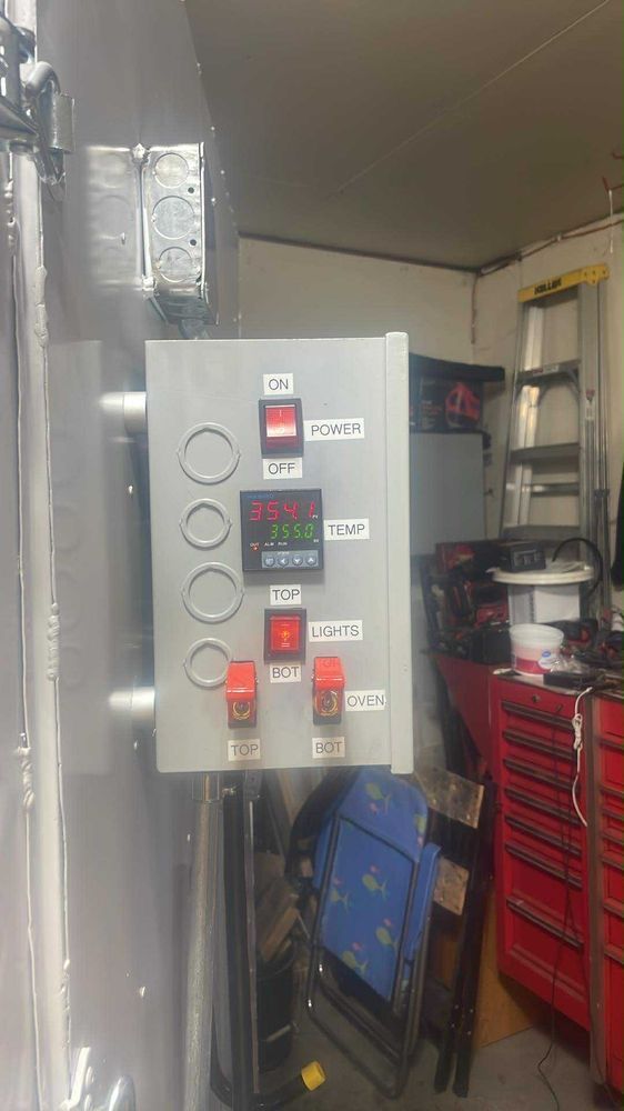 A gray electrical control box mounted on a wall in a workshop, featuring a digital temperature display and various switches.