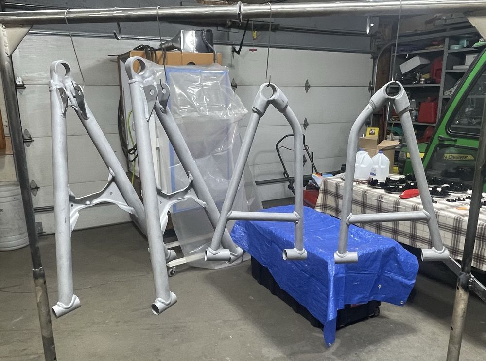 Four silver A-arm suspension components hang from a metal frame inside a garage workshop.