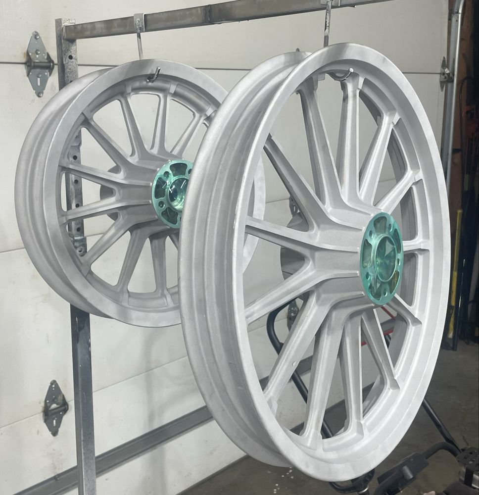 Two silver motorcycle wheels with teal center hubs hang from a metal rack in a garage.