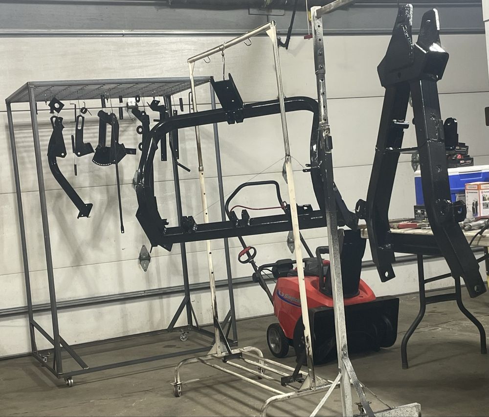 Various black-painted automotive metal parts hang from racks inside a garage, with a red snowblower in the background.