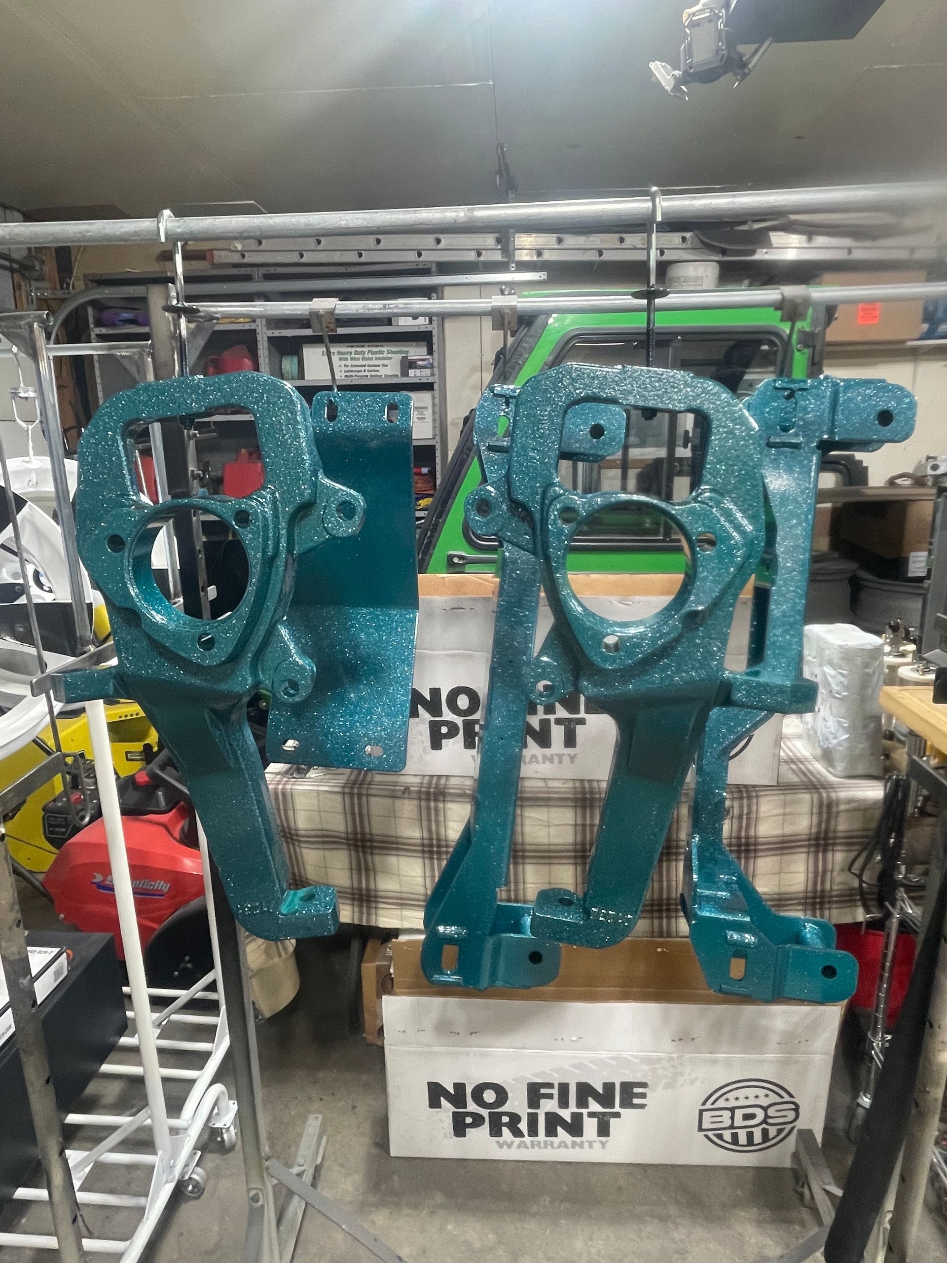 Two teal, powder-coated suspension steering knuckles hanging from a rack in a garage, with boxes in the background.
