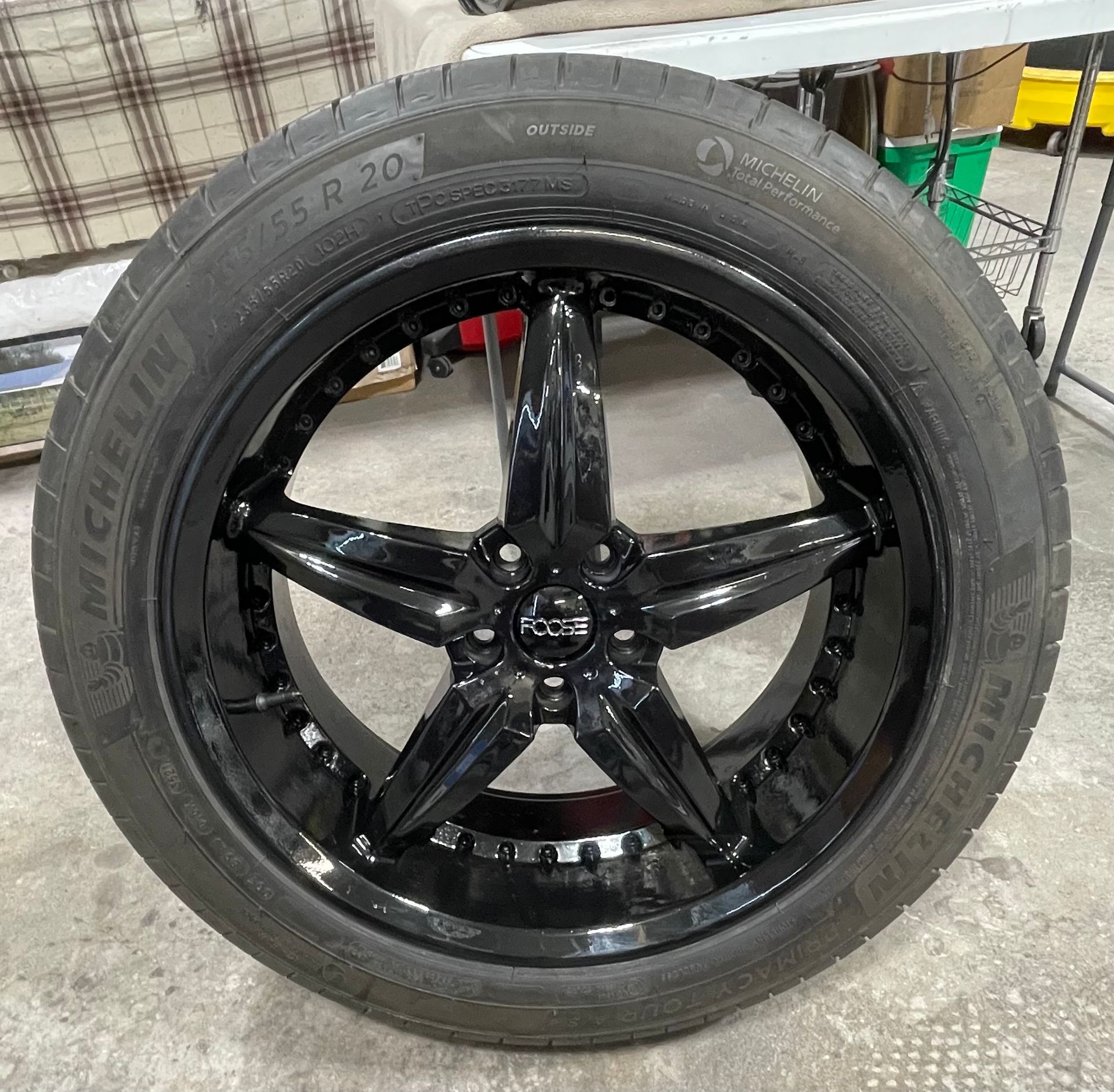 A glossy black five-spoke wheel with a Michelin tire mounted on a concrete floor in a garage setting.