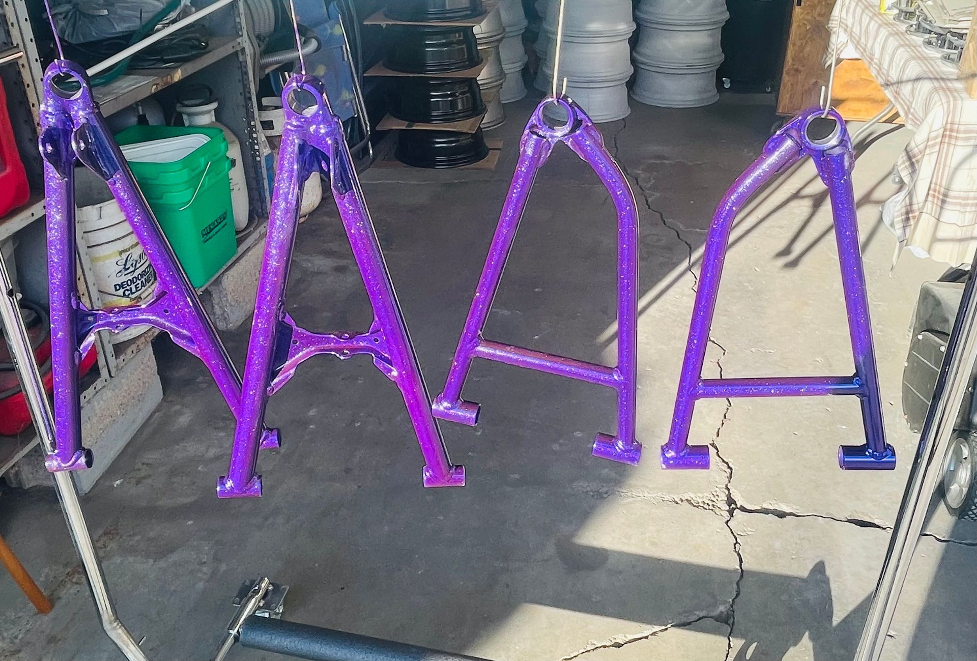 Four bright purple metal suspension A-arms hang in a row from hooks in an industrial workshop.