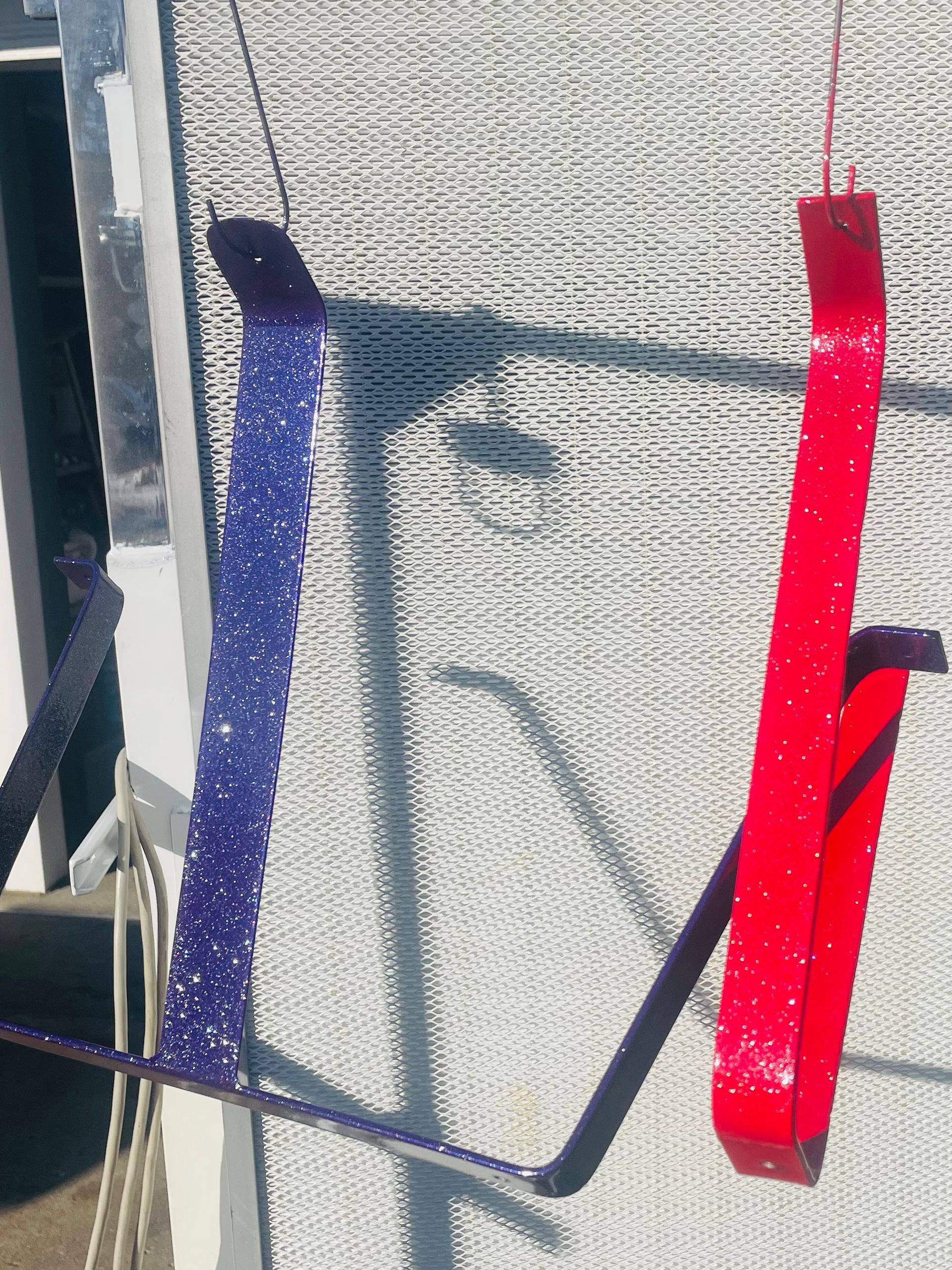 Two glittery, L-shaped metal brackets, one blue and one red, hang against a white mesh screen.
