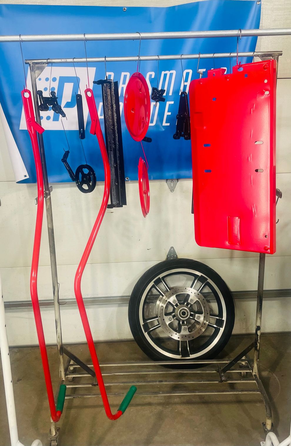 A metal rack holds various red and black automotive parts, including panels, rods, and a wheel, for powder coating.
