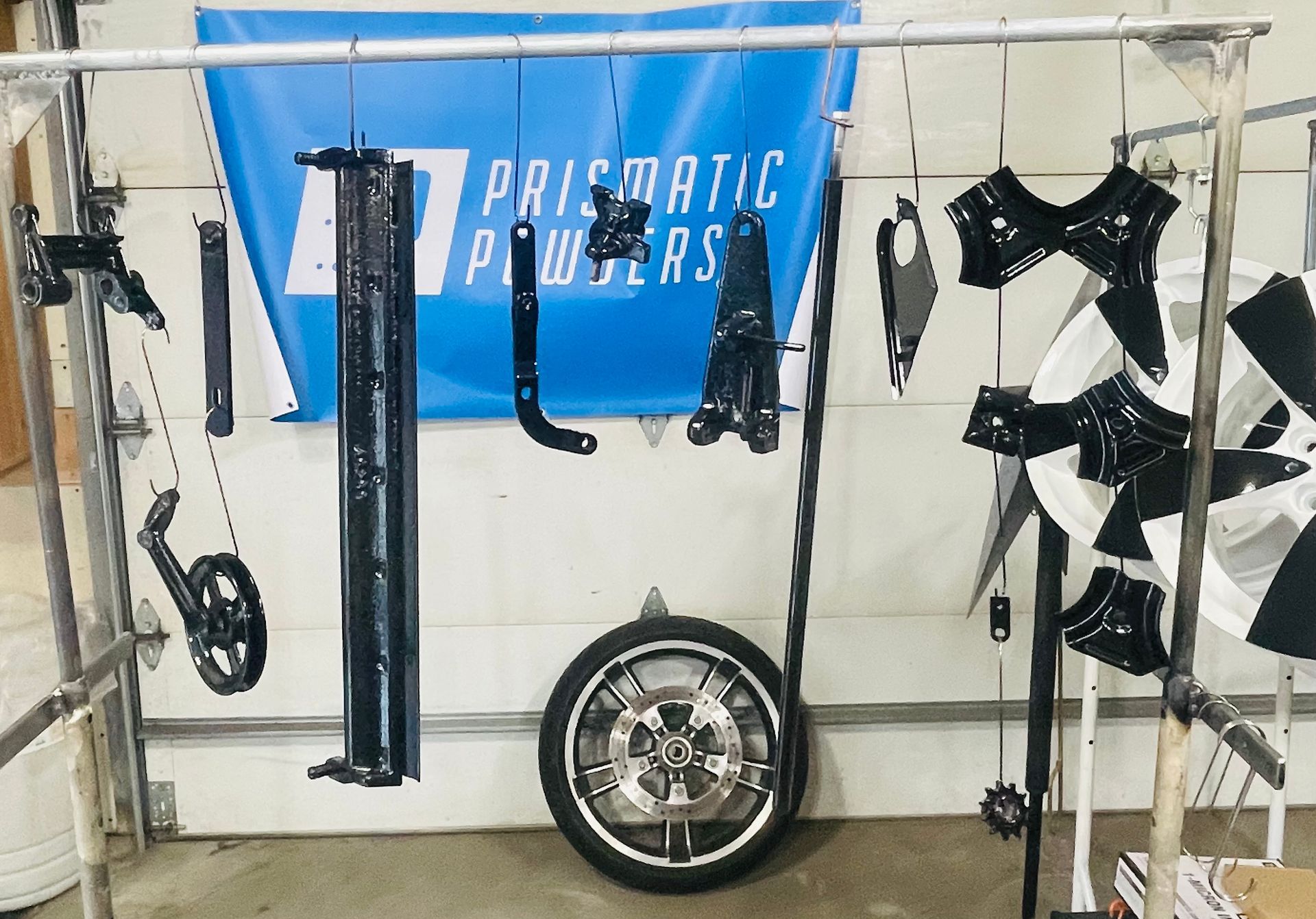 Black metal parts and a wheel hanging from a rack in a workshop, in front of a blue Prismatic Powders banner.