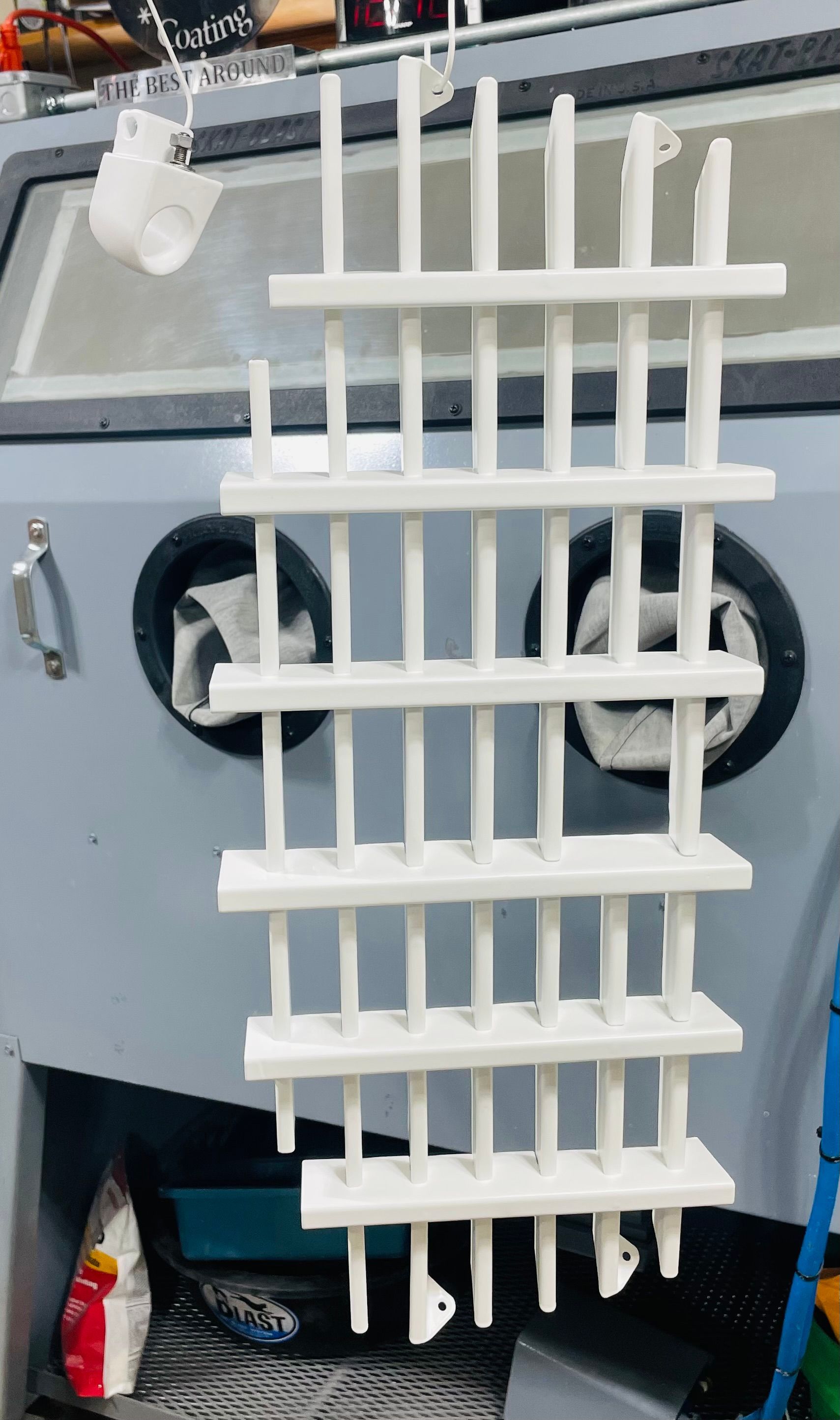 White rack with horizontal shelves and vertical bars hanging in front of an industrial sandblasting cabinet.
