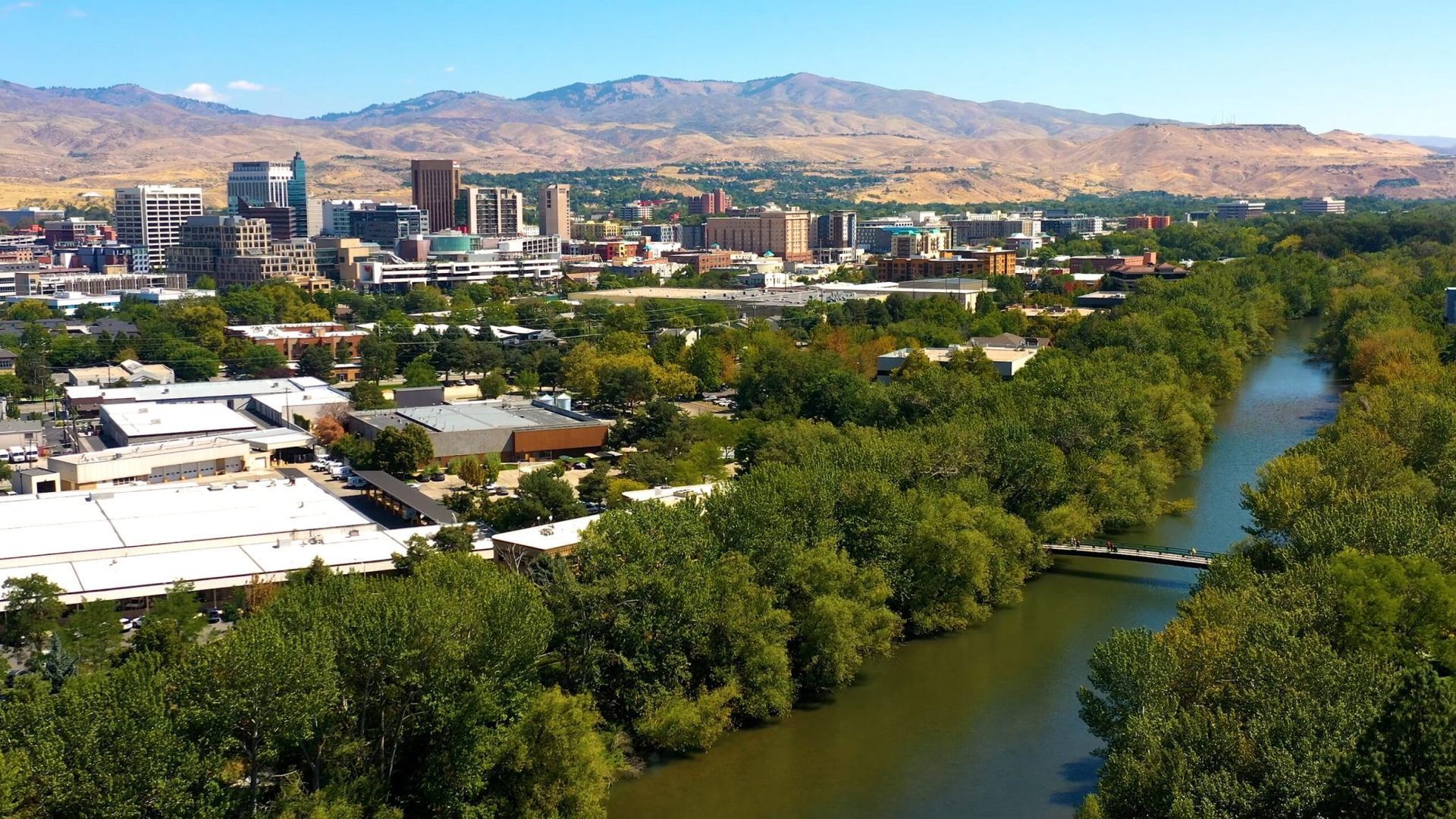 City skyline beside a green river with tree-lined banks and dry mountains in the distance