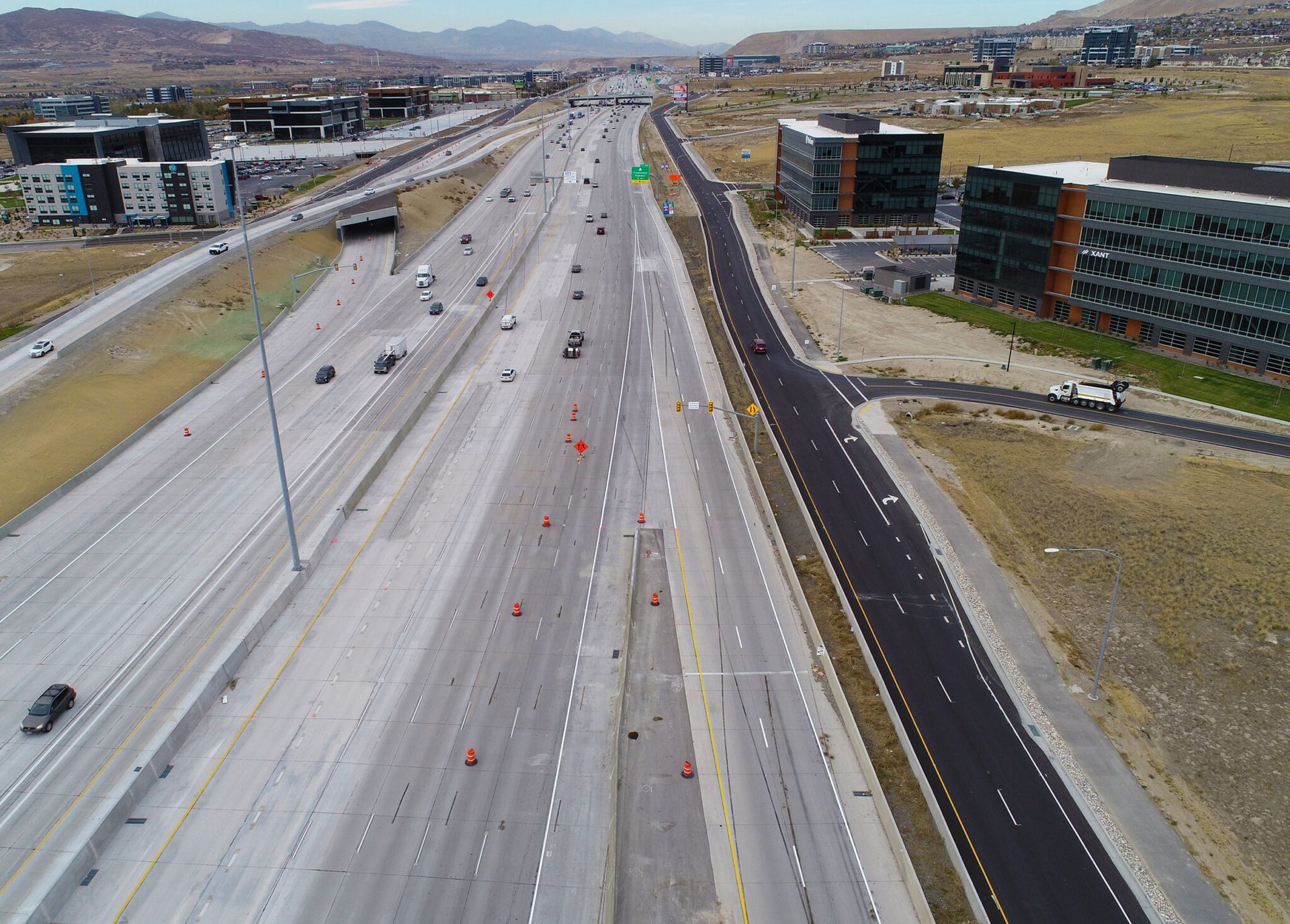Aerial view of a wide highway under construction with cones and sparse traffic, flanked by buildings and hills.