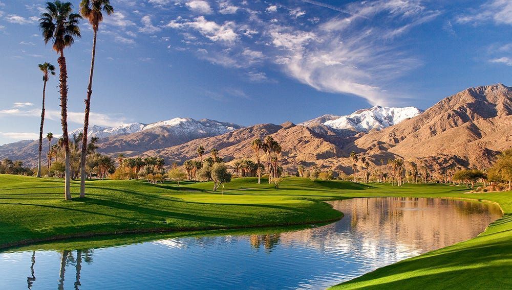 Desert golf course with palm trees, a pond, and snow-capped mountains under a blue sky