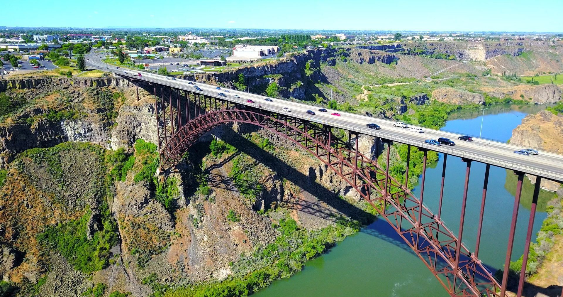 A long bridge spans a deep river gorge beside green hills and a small town under a bright sky.