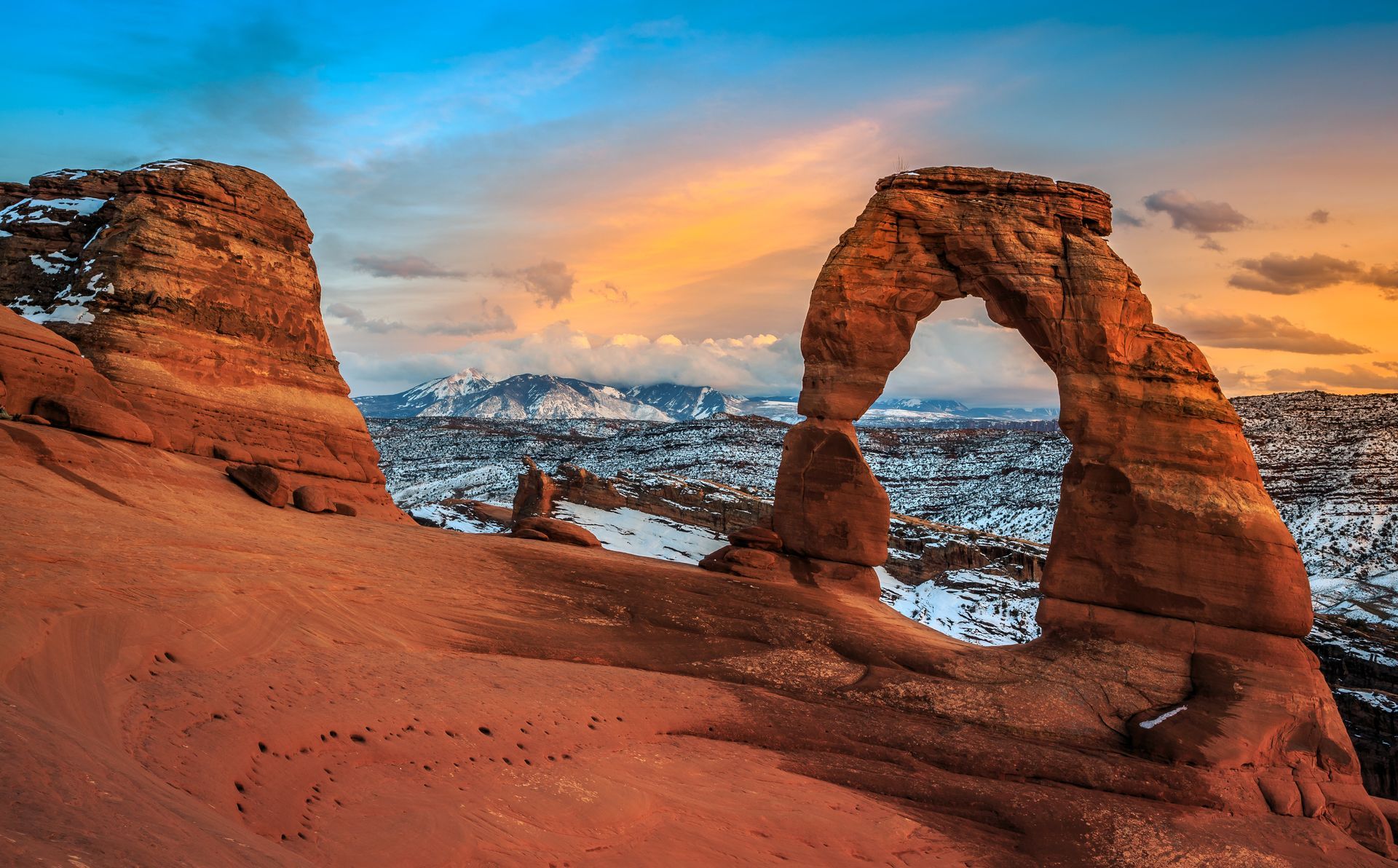 Red sandstone arch at sunset overlooking snowy mountains and a desert landscape