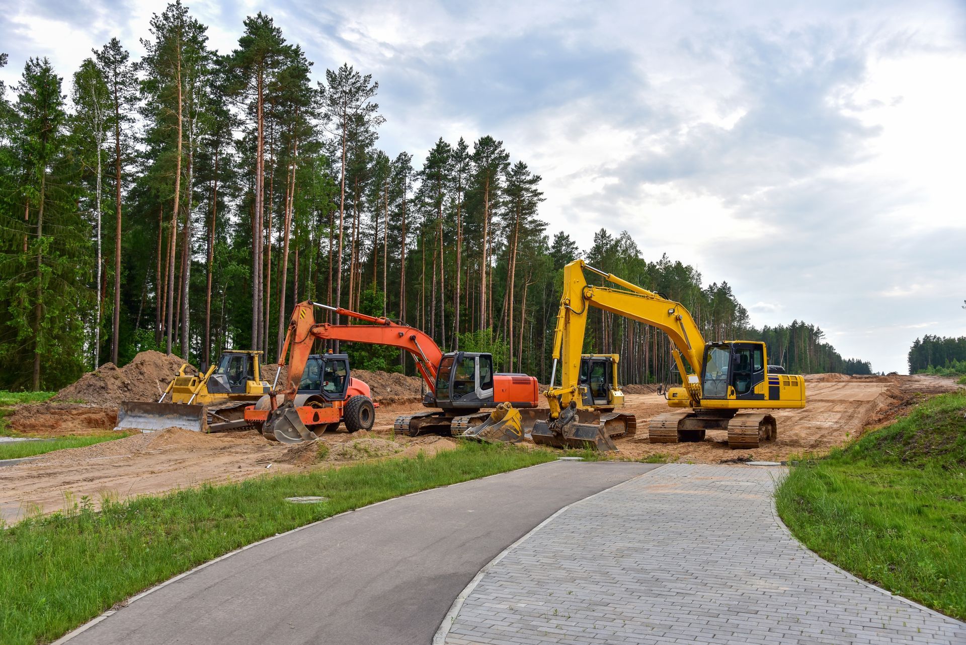 Construction site with orange and yellow excavators on a dirt road beside a forest path