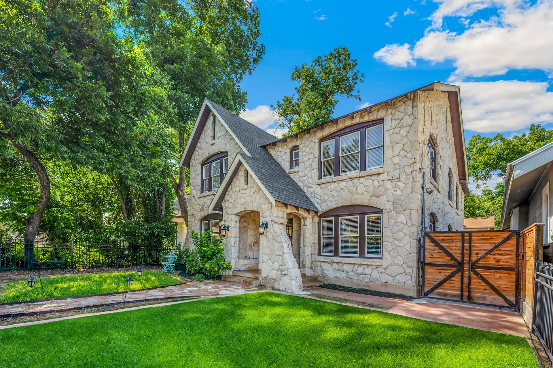 Stone two-story house with dark roof, front lawn, trees, and wooden gate under a bright blue sky