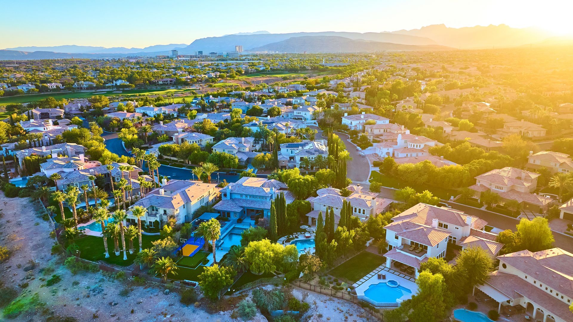 Sunlit aerial view of a suburban neighborhood with houses, trees, and distant mountains at sunset
