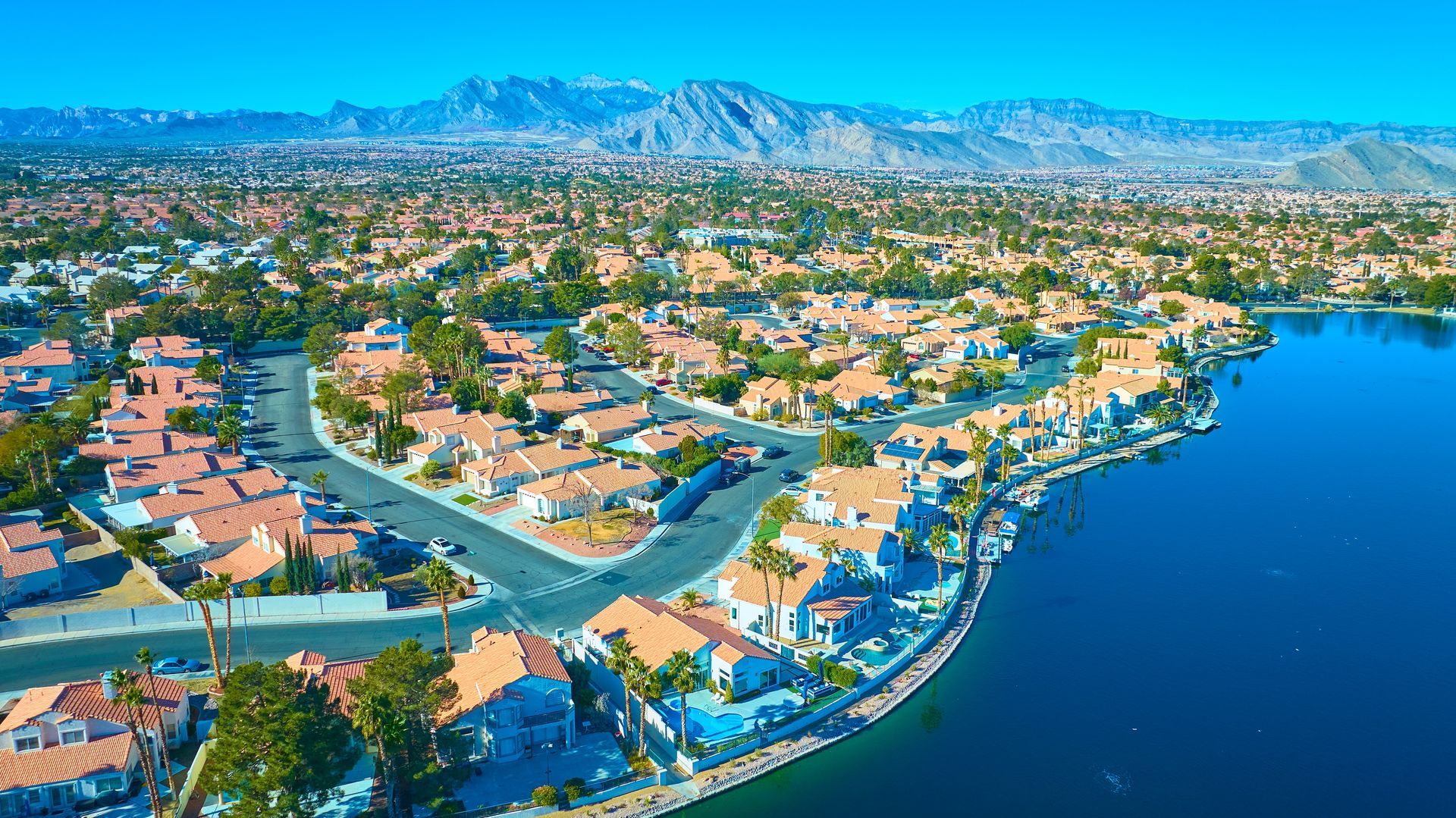 Aerial view of a suburban lakeside neighborhood with blue water and distant mountains under a clear sky