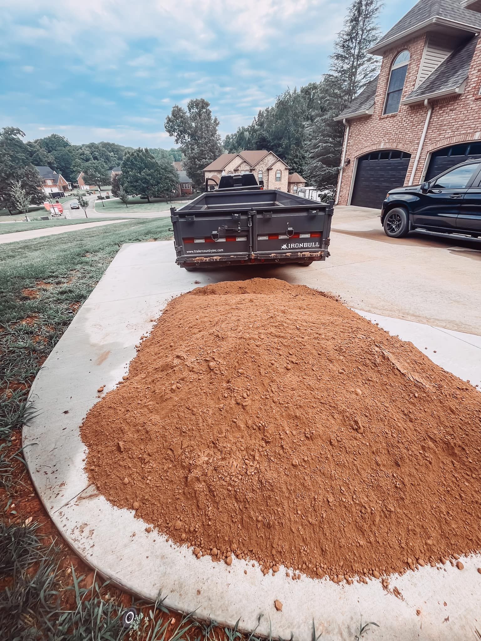 Dump truck unloading gravel on a curved driveway beside a brick house and parked SUV.