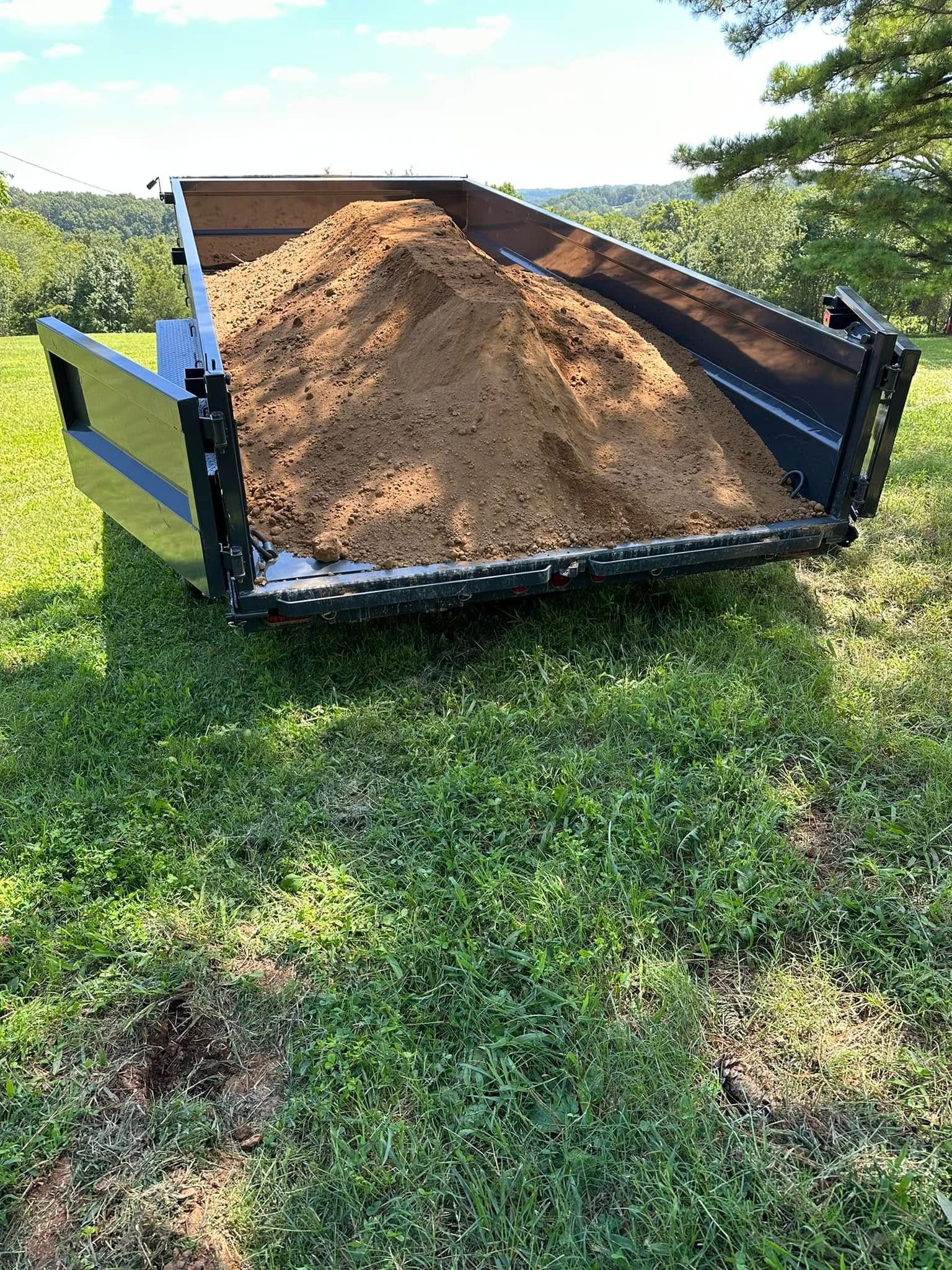 Black trailer loaded with a large mound of brown dirt on a grassy field in sunlight