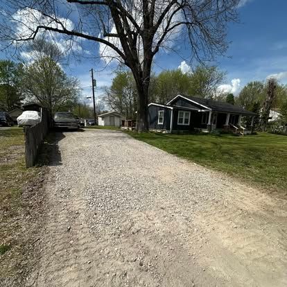 Gravel driveway leading to a small house under a large tree on a sunny day